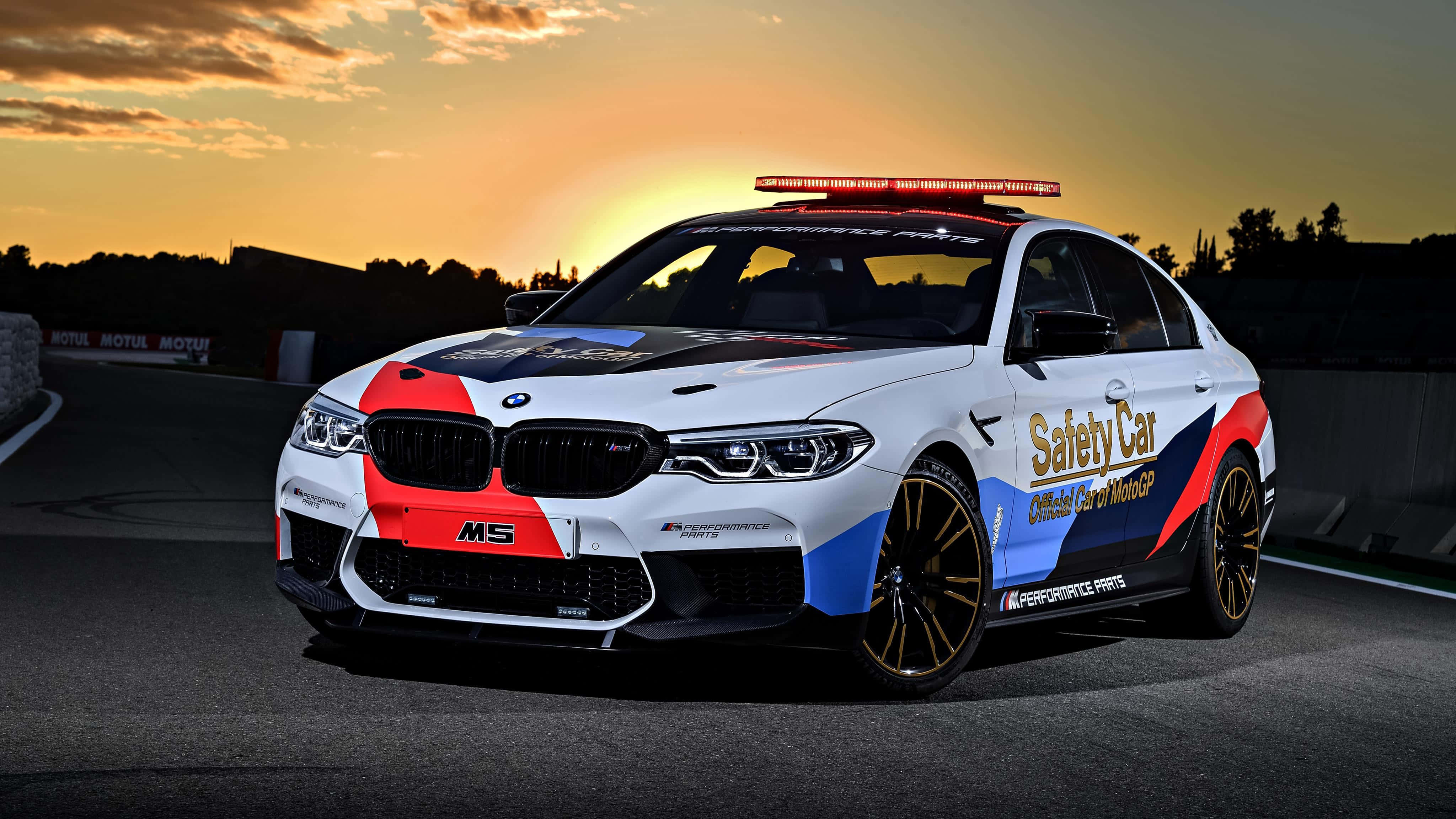 Safety Car 桌布 壁紙 — 一輛紅藍白塗裝的BMW M5安全車在日落時分，伴隨著閃爍燈光和溫暖天空，在賽道上奔馳。
