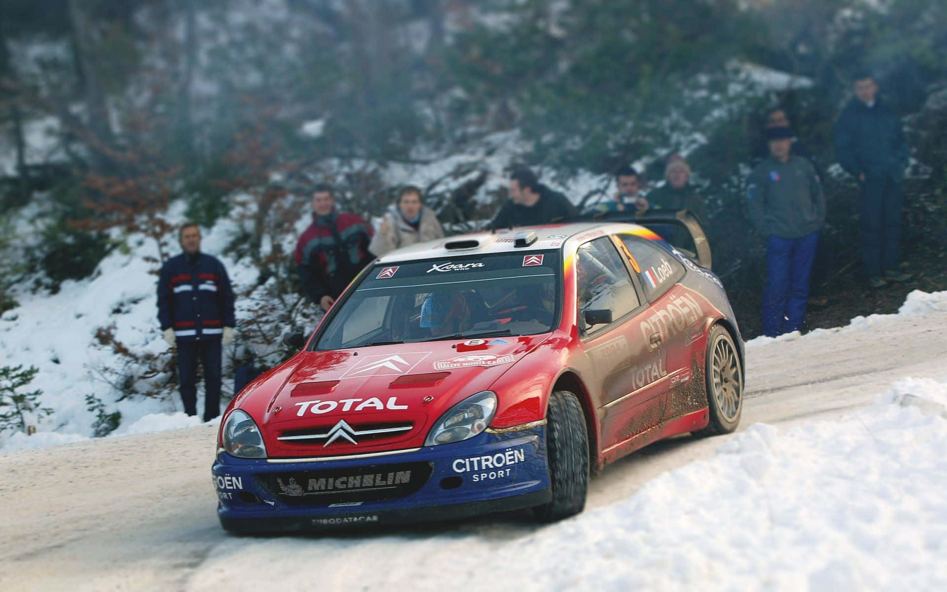 Ice Racing 桌布 壁紙 — 一輛紅藍色的雪鐵龍拉力賽車在白雪覆蓋的森林賽道上飛馳，觀眾在旁觀看。