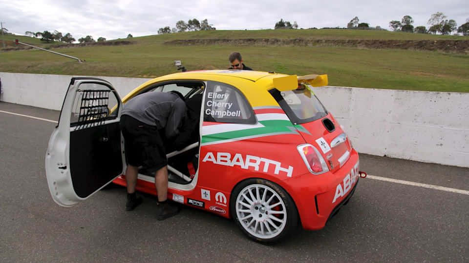 Abarth 695 桌布 壁紙 — 一輛紅黃綠色的 Abarth 賽車在賽道上打開車門，一個人正探身進入。