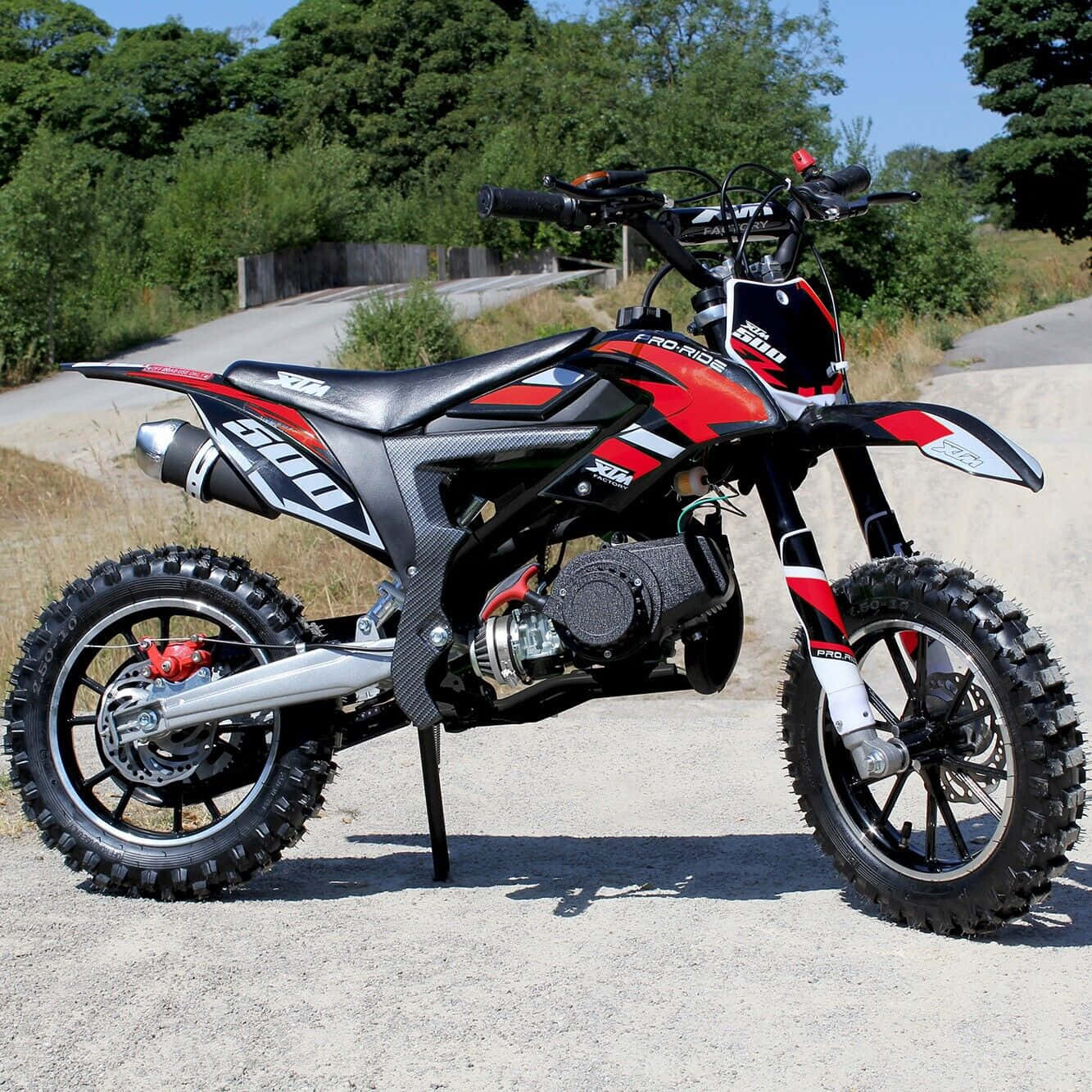 Dirtbike 桌布 壁紙 — 一輛紅黑白色的越野機車，配備防滑輪胎，停在碎石路上，周圍綠樹成蔭，天空清澈湛藍。