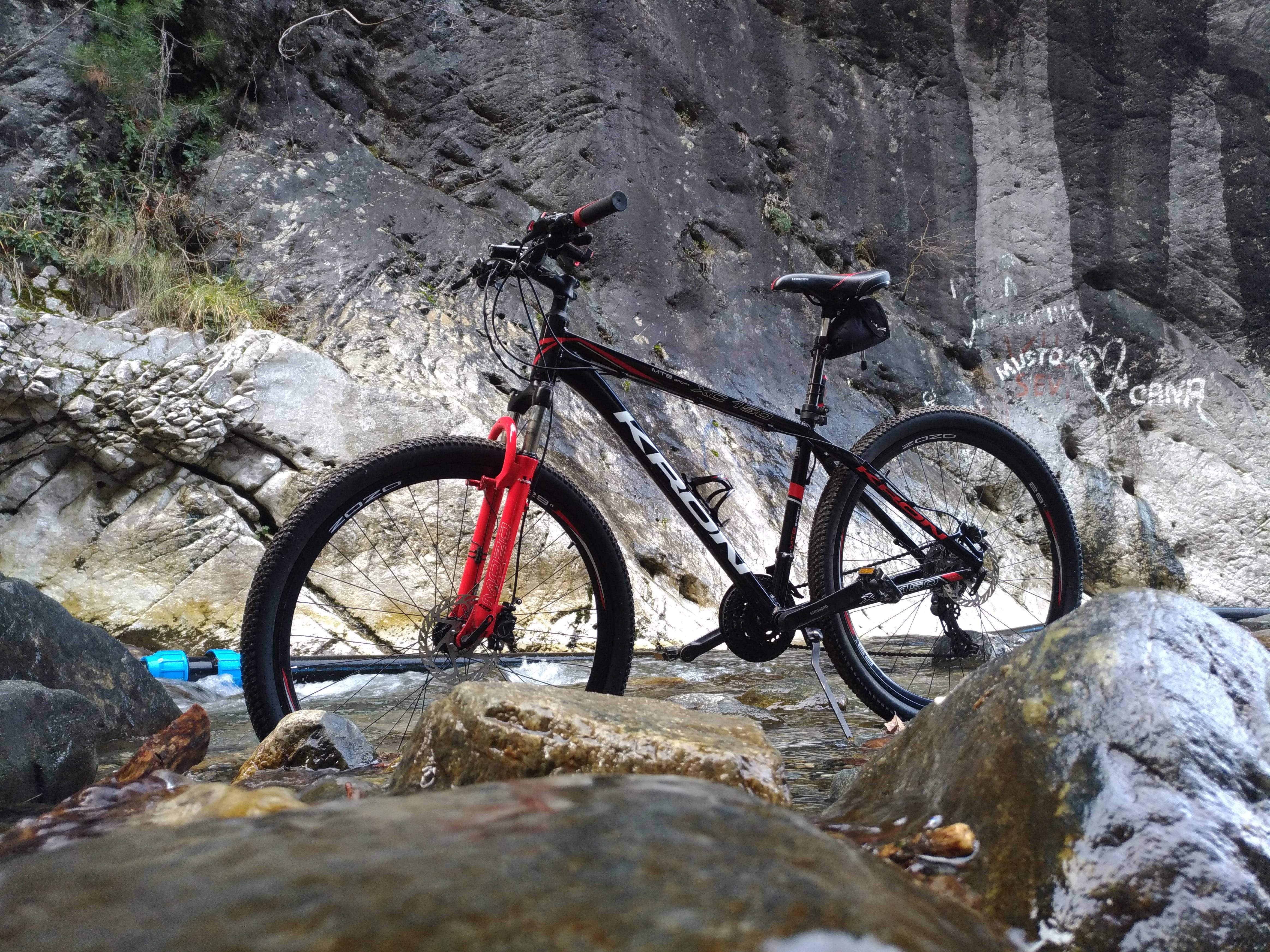Mountain Bike 桌布 壁紙 — 一輛紅黑色登山車佇立在淺山溪中，四周是濕石和崎嶇懸崖。