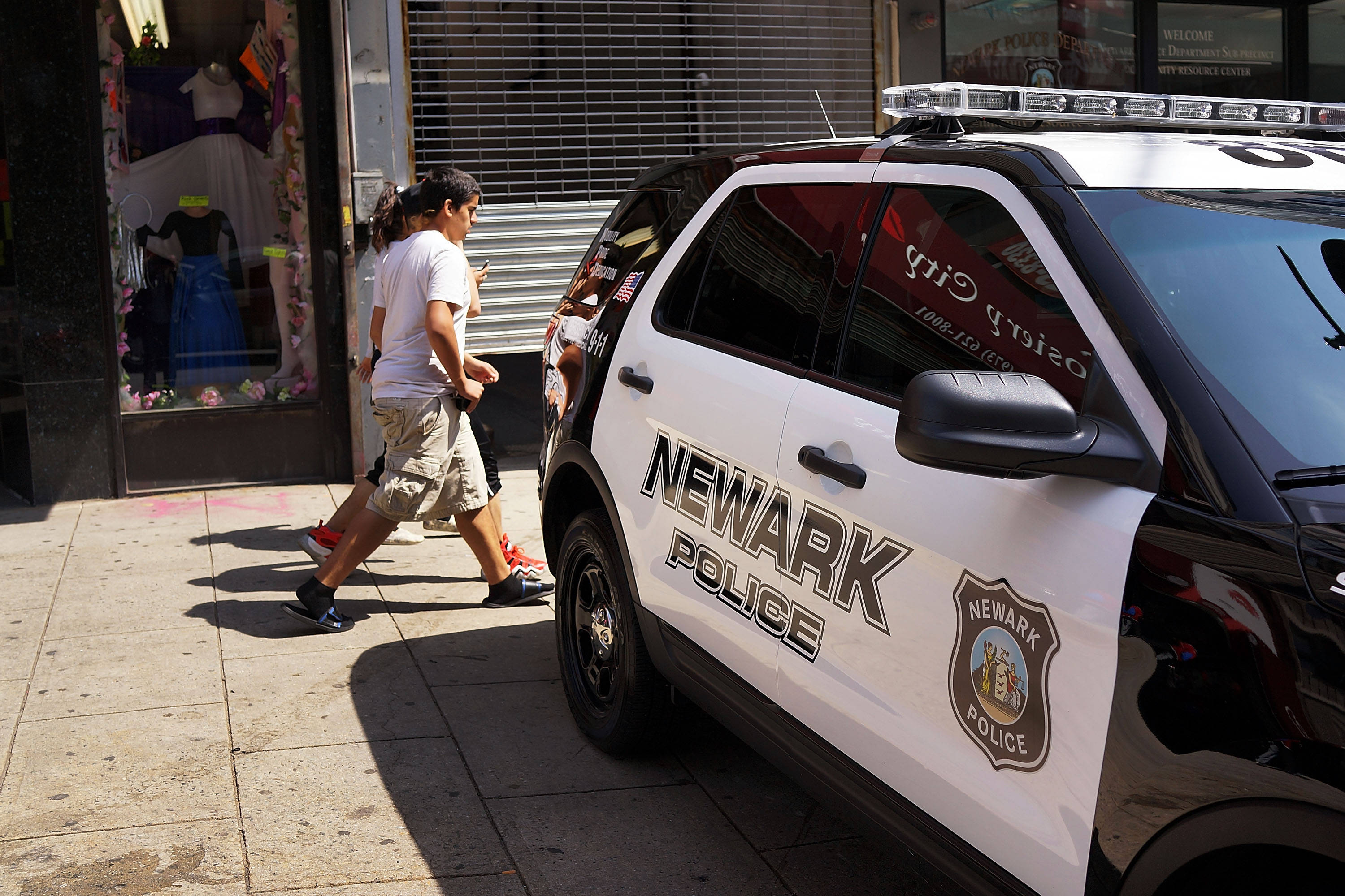 警察 桌布 壁紙 — 一輛紐瓦克警車停放在陽光明媚的城市人行道上，行人正走過一間精品店門面。