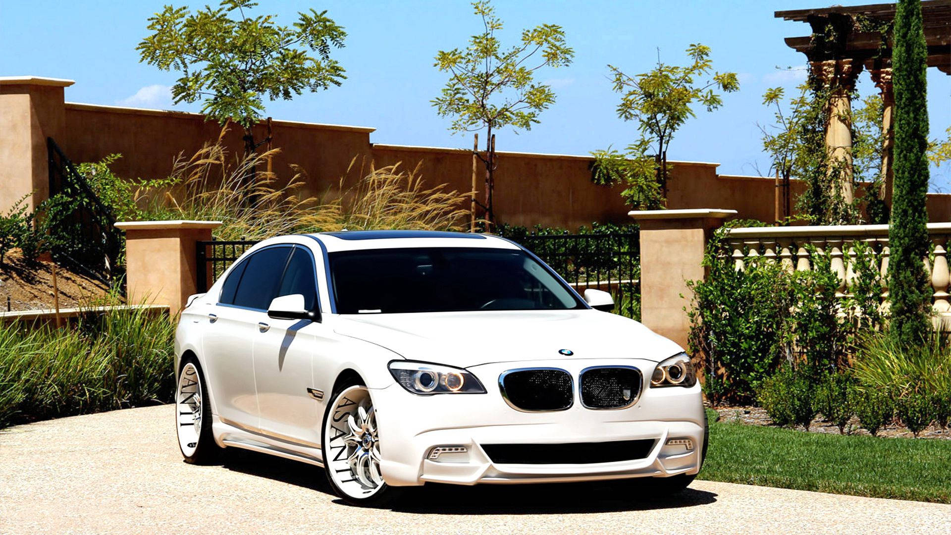 BMW 動圖 桌布 壁紙 — 一輛純白色BMW轎車配備定制輪圈，在陽光下閃耀，停在碎石車道上，旁邊是灰泥牆和修剪整齊的花園。