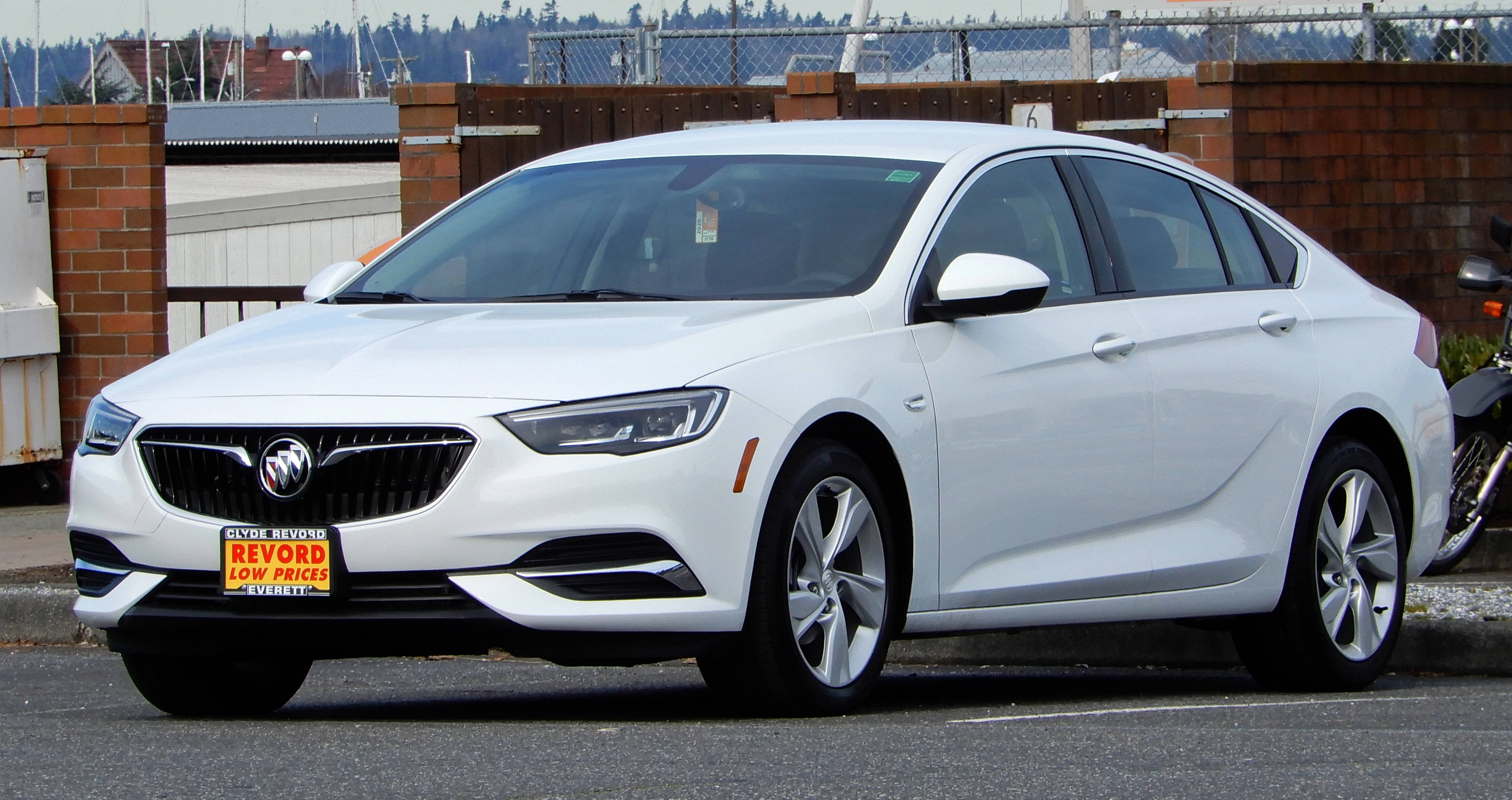 Buick 桌布 壁紙 — 一輛純白色別克轎車停在瀝青停車場，周圍有磚牆和遠處的樹木，天空多雲。