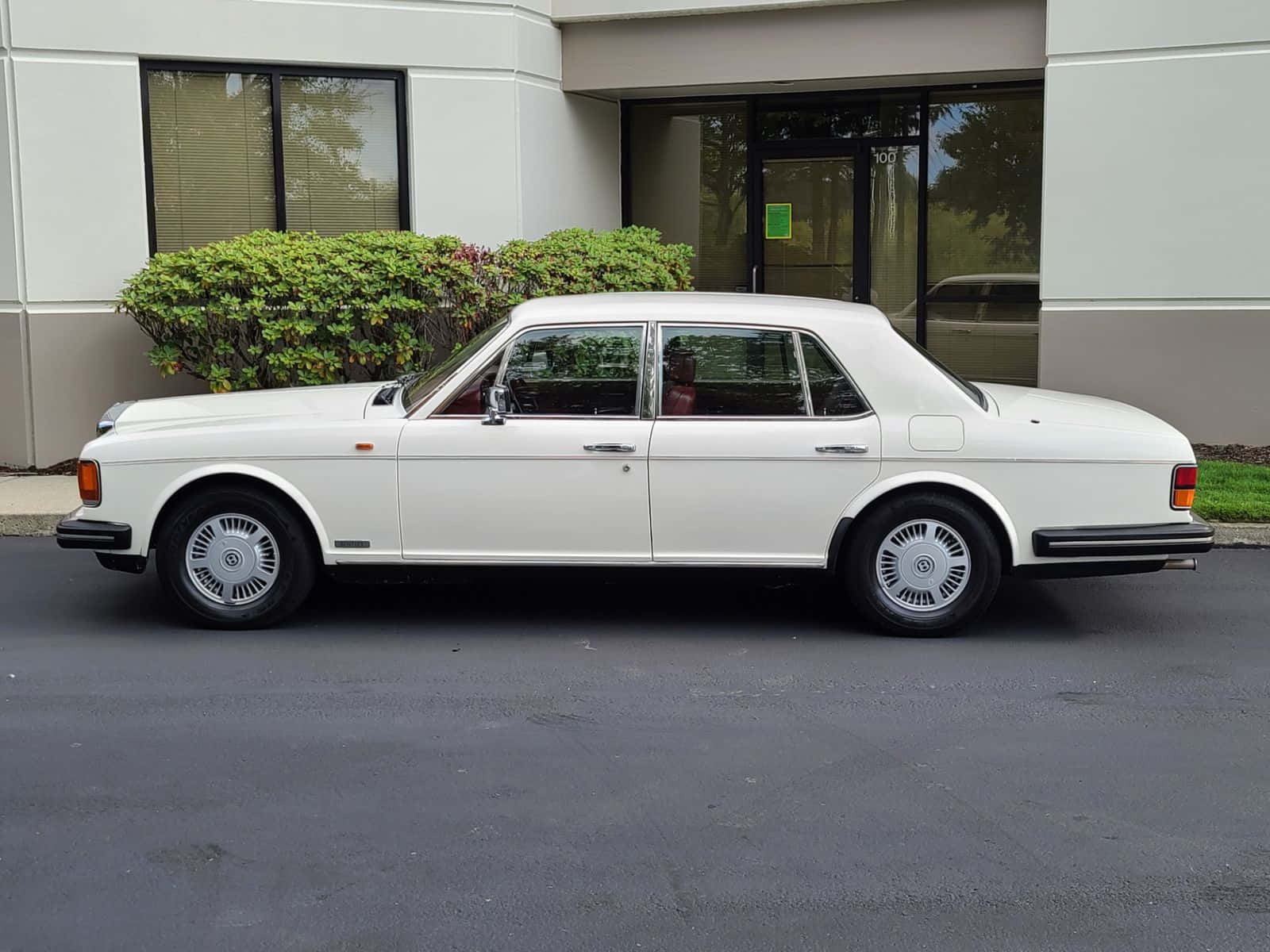 Bentley Eight 桌布 壁紙 — 一輛純白色勞斯萊斯 Silver Shadow II 停放在現代建築物前，周圍有綠色灌木。