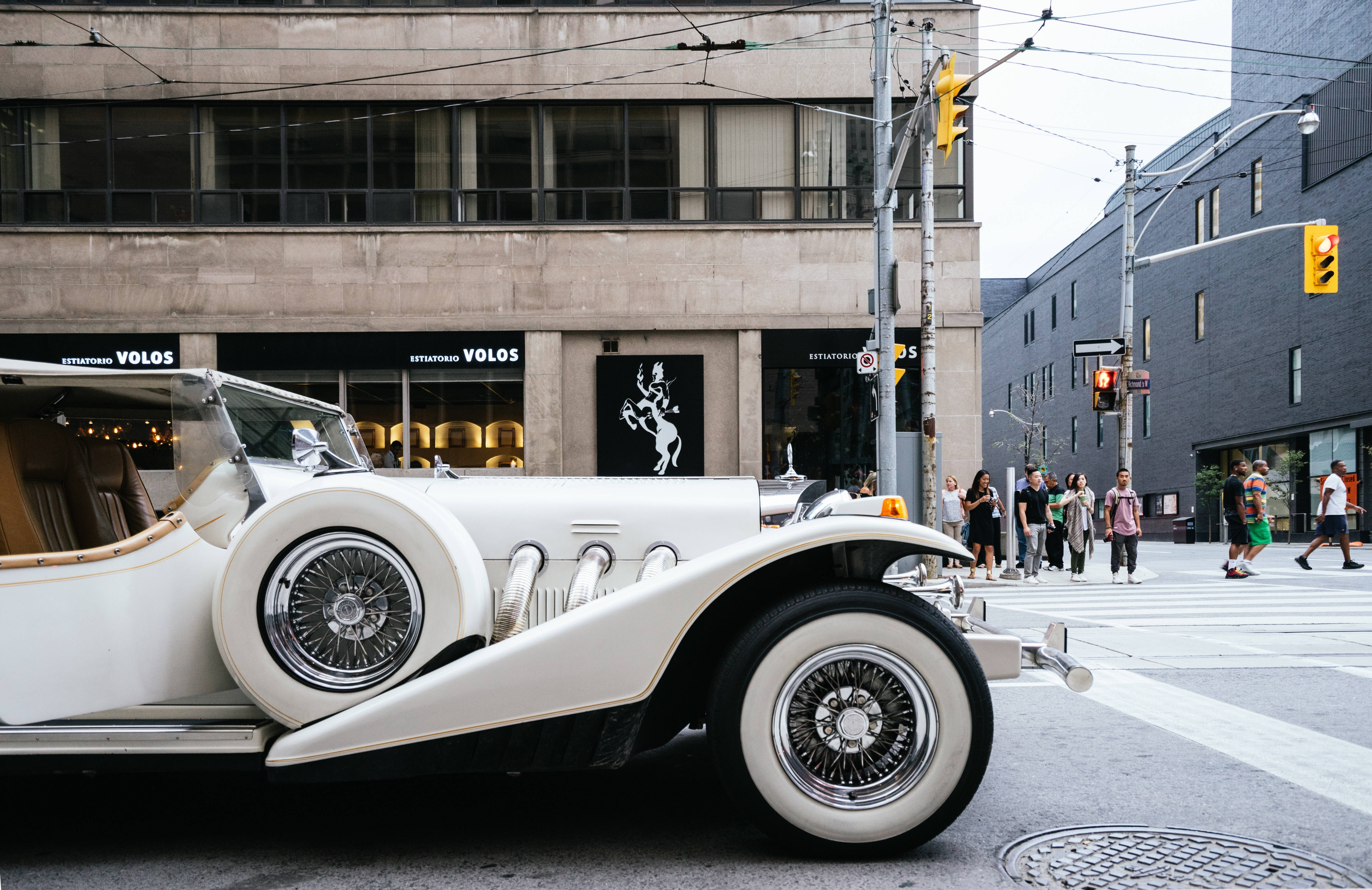 Cool Car 桌布 壁紙 — 一輛純白色復古敞篷車配白色輪胎停放在城市行人穿越道，行人正走過現代建築。