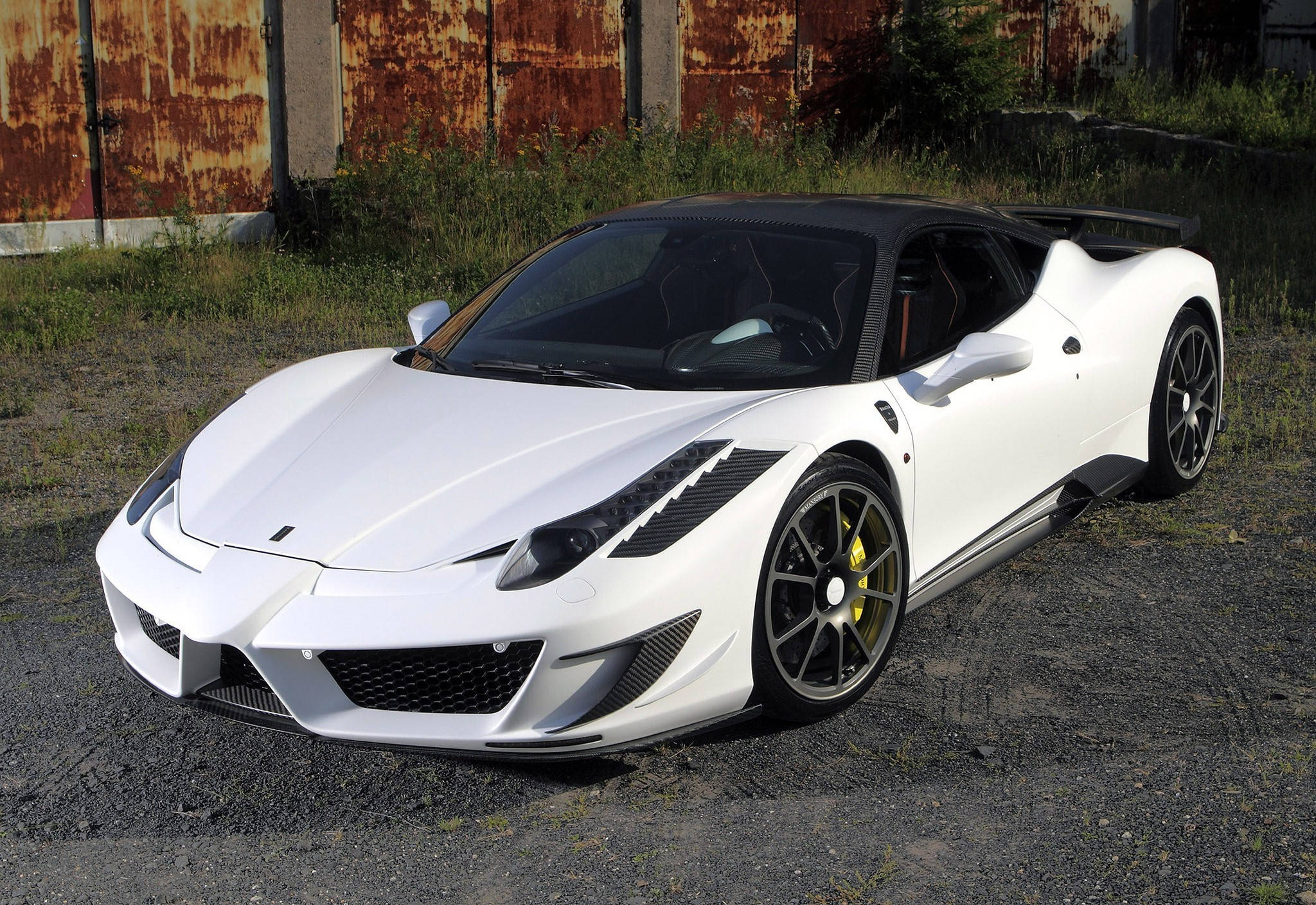white ferrari 桌布 壁紙 — 一輛純白色法拉利跑車，帶有黑色飾邊和黃色制動卡鉗，停在碎石路上，旁邊是生鏽的金屬牆。