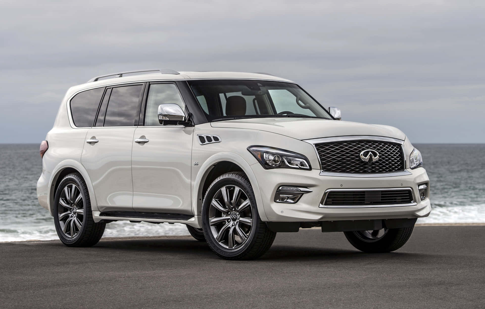 Infiniti Qx80 桌布 壁紙 — 一輛純白色的 Infiniti QX80 SUV 停泊在海岸公路上，背後有海浪拍打，天空多雲。