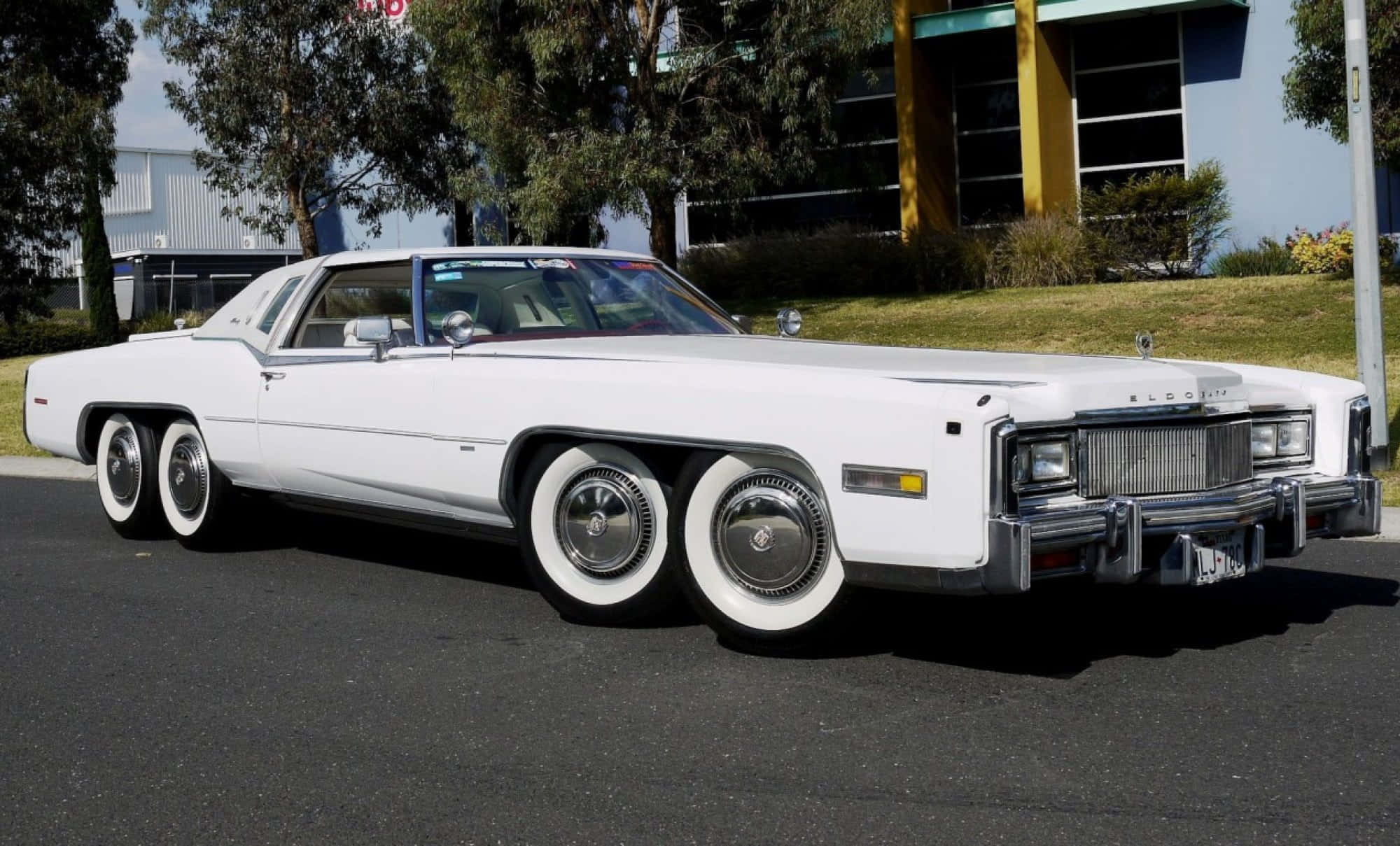 Cadillac Eldorado 桌布 壁紙 — 一輛純白色的1970年代凱迪拉克，配備三組後輪，在明亮的陽光下閃閃發光，停放在靠近樹木和現代建築的柏油路面上。