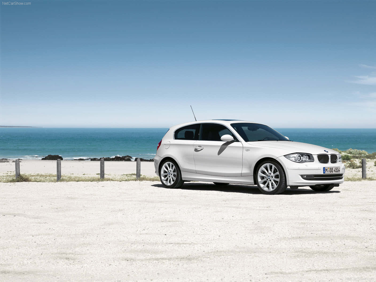 Bmw 1 Series 桌布 壁紙 — 一輛純白色的BMW 1系列掀背車停放在陽光普照的海岸碎石停車場，背景是海洋和天空。
