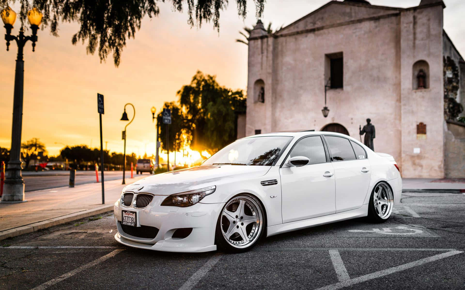 Tuned Car 桌布 壁紙 — 一輛純白色的BMW房車在停車場中閃耀，夕陽映襯著歷史建築和溫暖的金色光芒。