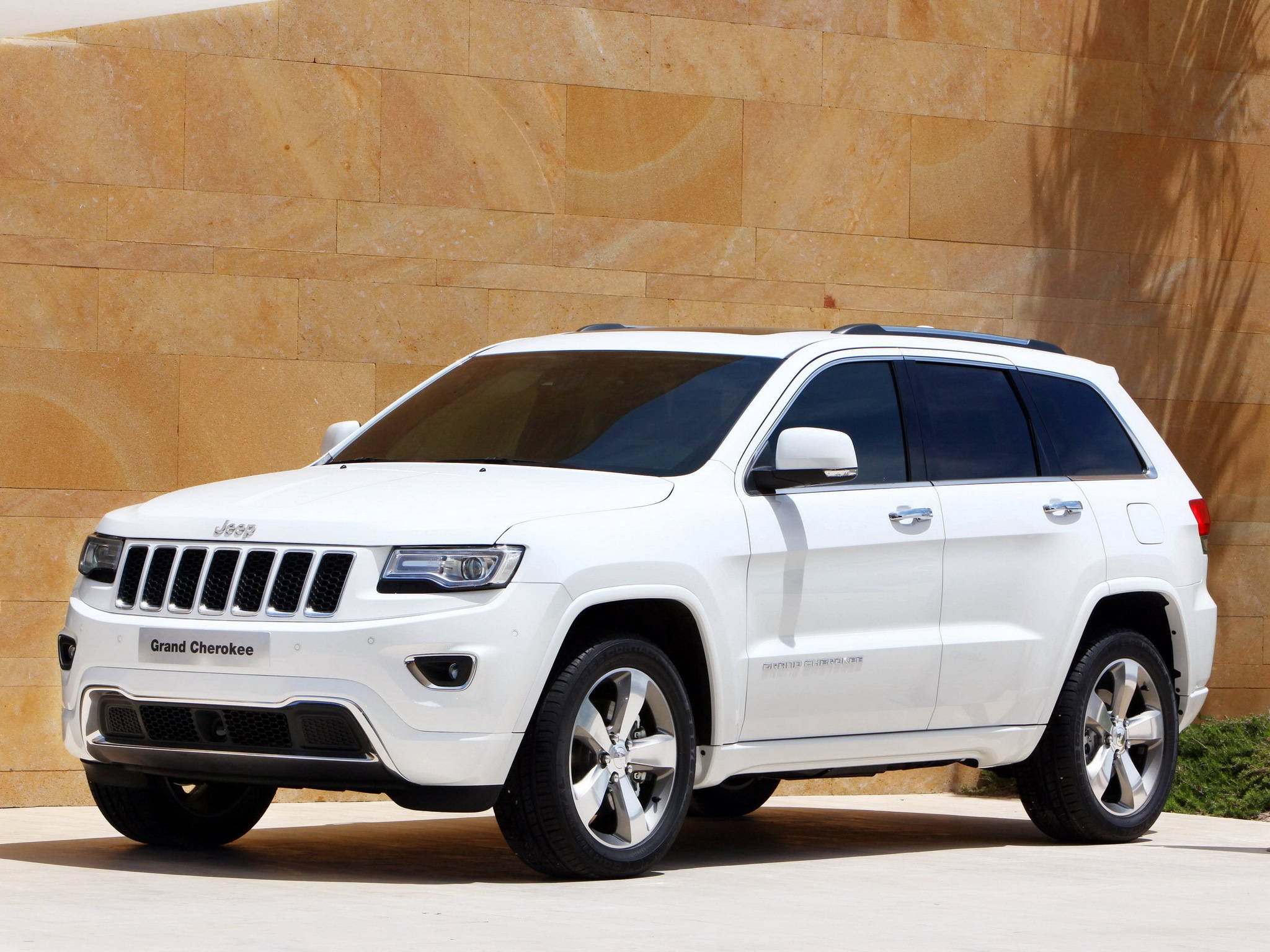 吉普 桌布 壁紙 — 一輛純白色的Jeep Grand Cherokee SUV在明亮的日光下停靠在溫暖的米色石牆旁。