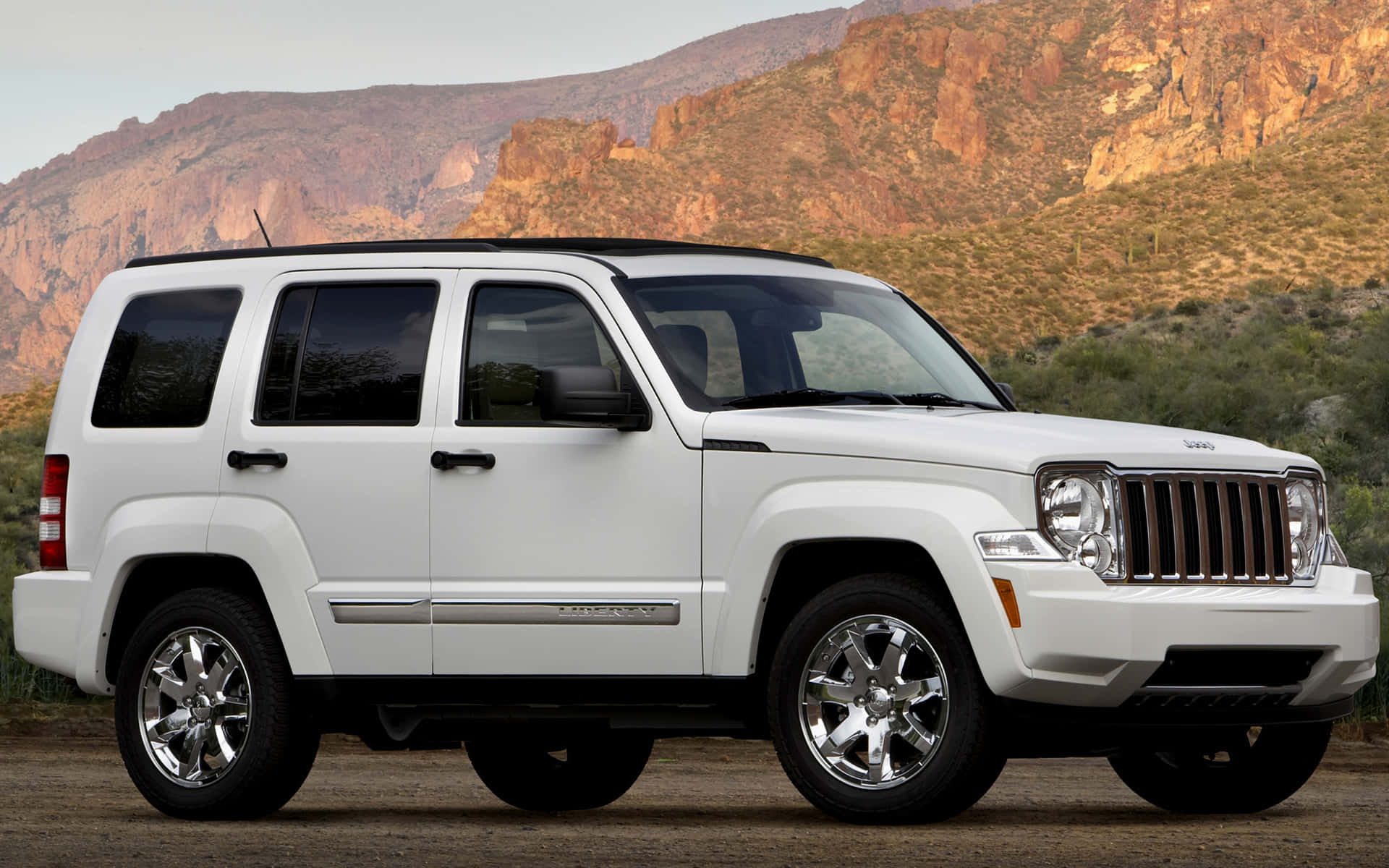 Jeep Liberty 桌布 壁紙 — 一輛純白色的Jeep Patriot SUV停泊在泥土上，背景是崎嶇的紅色岩石山脈和稀疏的沙漠植被，天空晴朗。