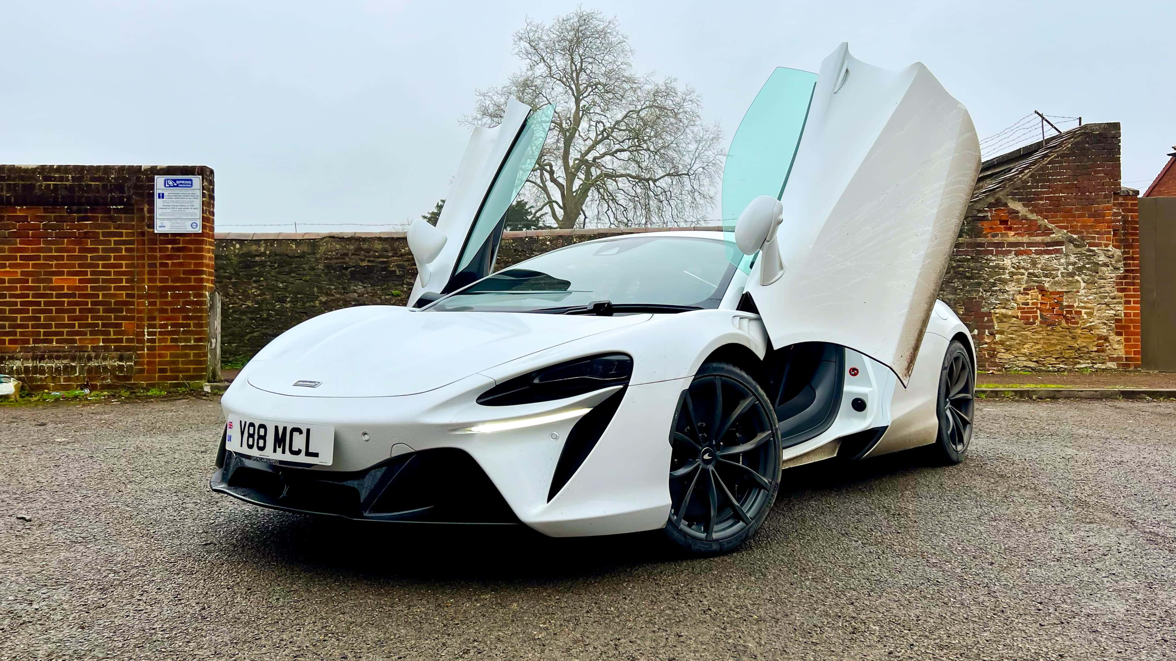 麥拉倫 桌布 壁紙 — 一輛純白色的McLaren跑車，鷗翼車門敞開，停在磚牆旁的礫石地上，天空多雲。