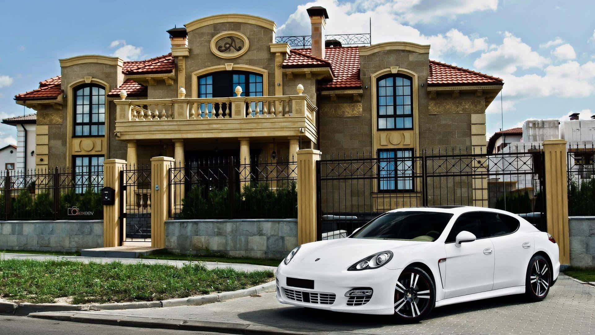 Beautiful House 桌布 壁紙 — 一輛純白色的保時捷Panamera停放在鋪砌的車道上，前方是一座宏偉的米色豪宅，擁有紅色瓦片屋頂和華麗的鐵門，在明亮的藍天下。
