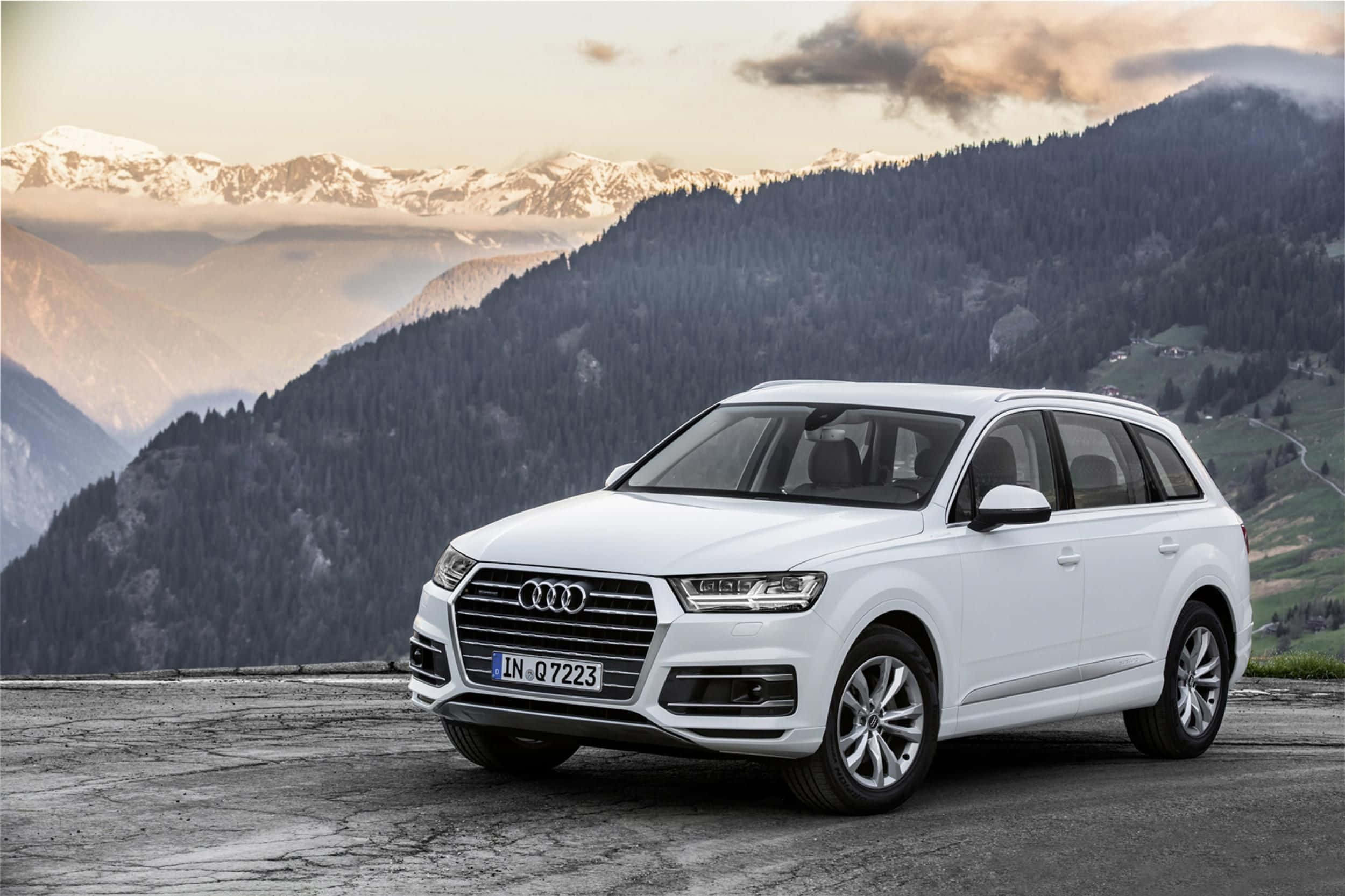 Audi Q7 桌布 壁紙 — 一輛純白色的奧迪Q7 SUV停在山頂觀景點，俯瞰著白雪覆蓋的山峰和森林覆蓋的山坡，天空呈現柔和的金色。