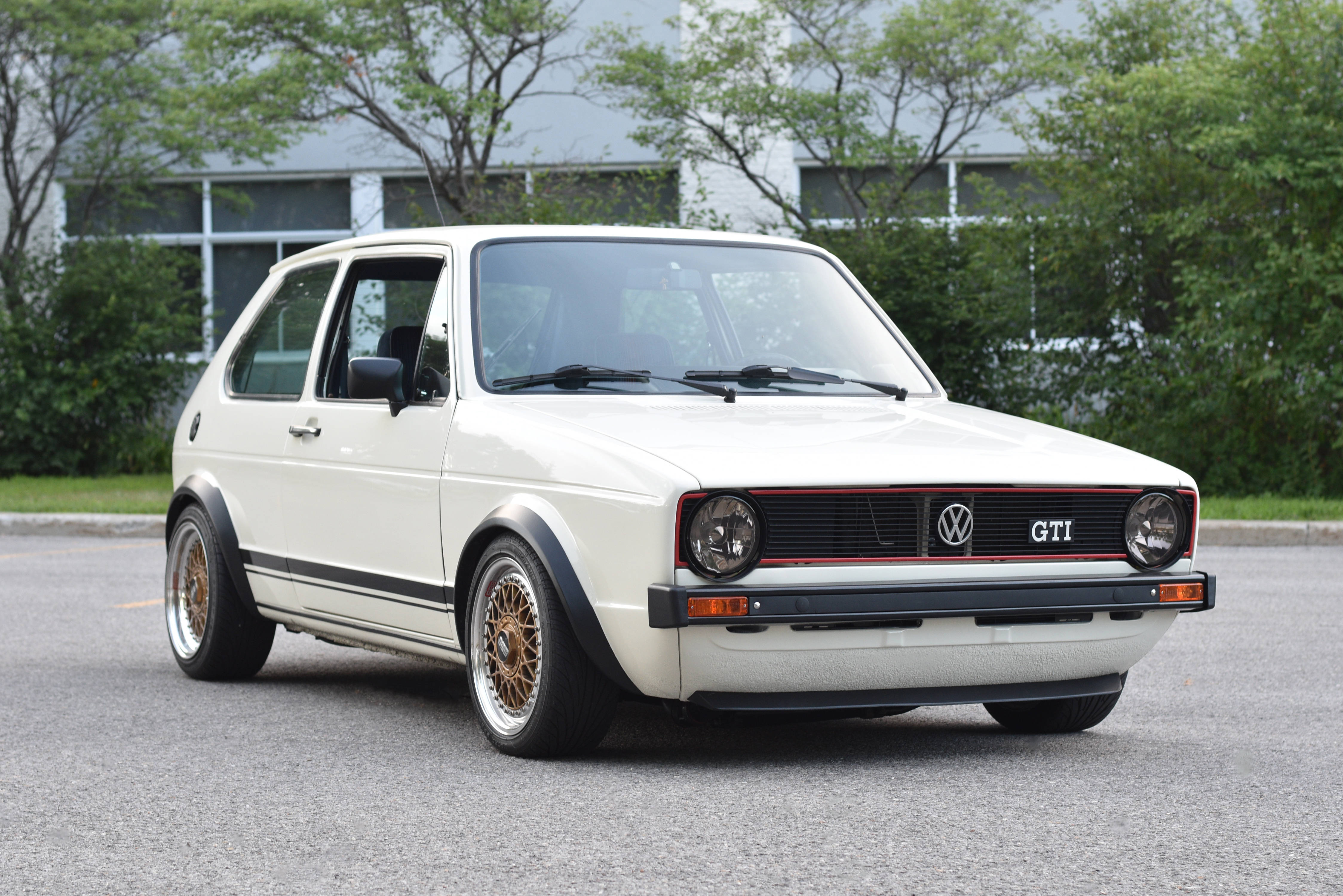 Gti 桌布 壁紙 — 一輛純白色的復古福斯Golf GTI，配金色輪圈，停在柏油路上，周圍有綠樹和現代建築。