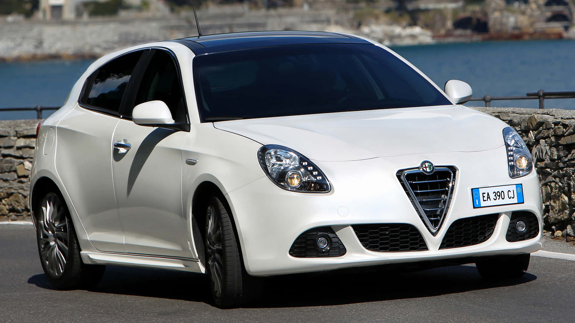 Alfa Romeo Giulietta 桌布 壁紙 — 一輛純白色的愛快羅密歐Giulietta停泊在沿海道路上，背後有石牆和藍色大海。