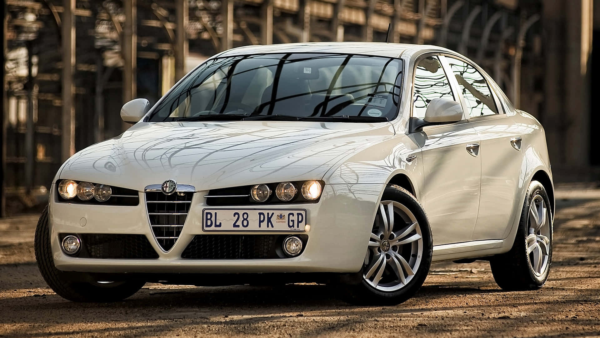 Alfa Romeo 159 桌布 壁紙 — 一輛純白色的愛快羅密歐轎車在溫暖陽光下閃閃發光，停泊在工業建築附近的塵土飛揚的地面上。