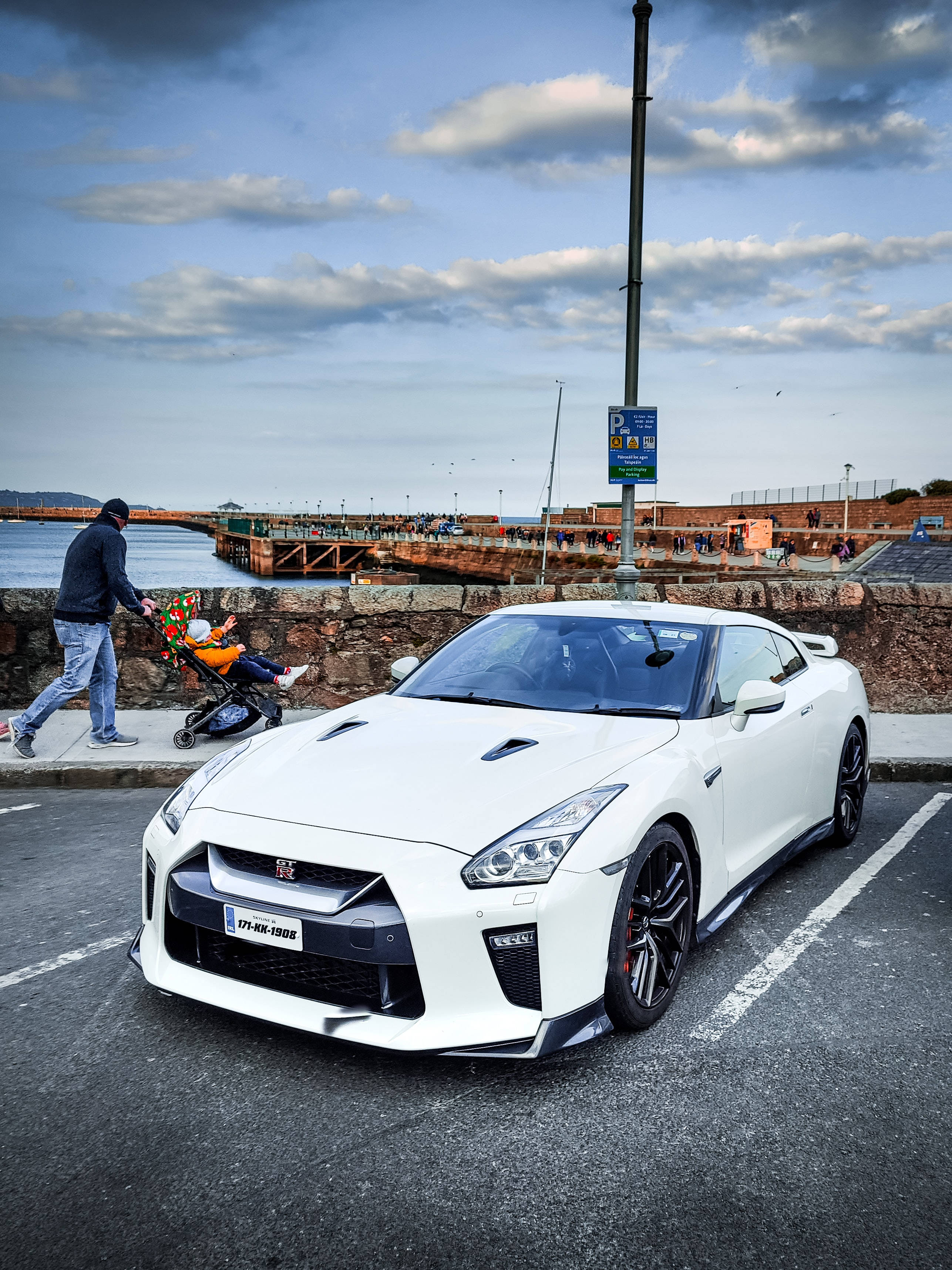 Gtr 桌布 壁紙 — 一輛純白色的日產GT-R停在海邊停車場，一名男子推著嬰兒車向水邊走去。