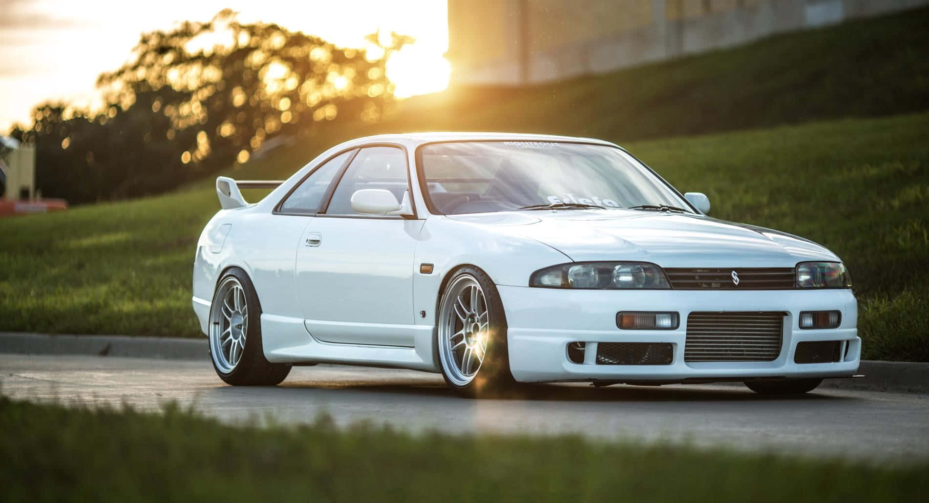 R33 GT-R 桌布 壁紙 — 一輛純白色的日產Skyline R32跑車在夕陽金光下閃耀，行駛在兩旁長滿青草的道路上。