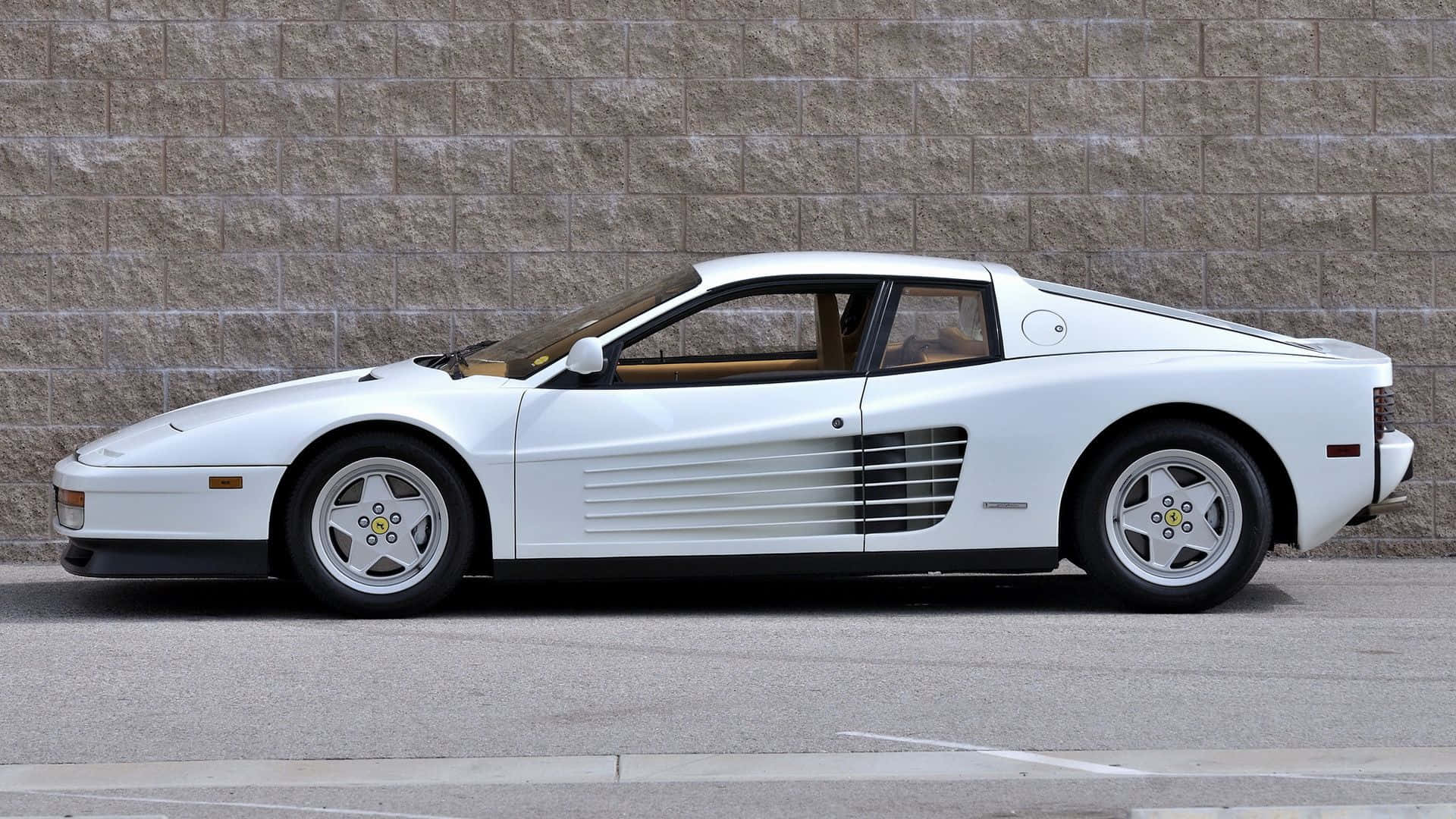 Ferrari Testarossa 桌布 壁紙 — 一輛純白色的法拉利 Testarossa 停放在鋪砌街道上、帶有紋理的石牆旁。