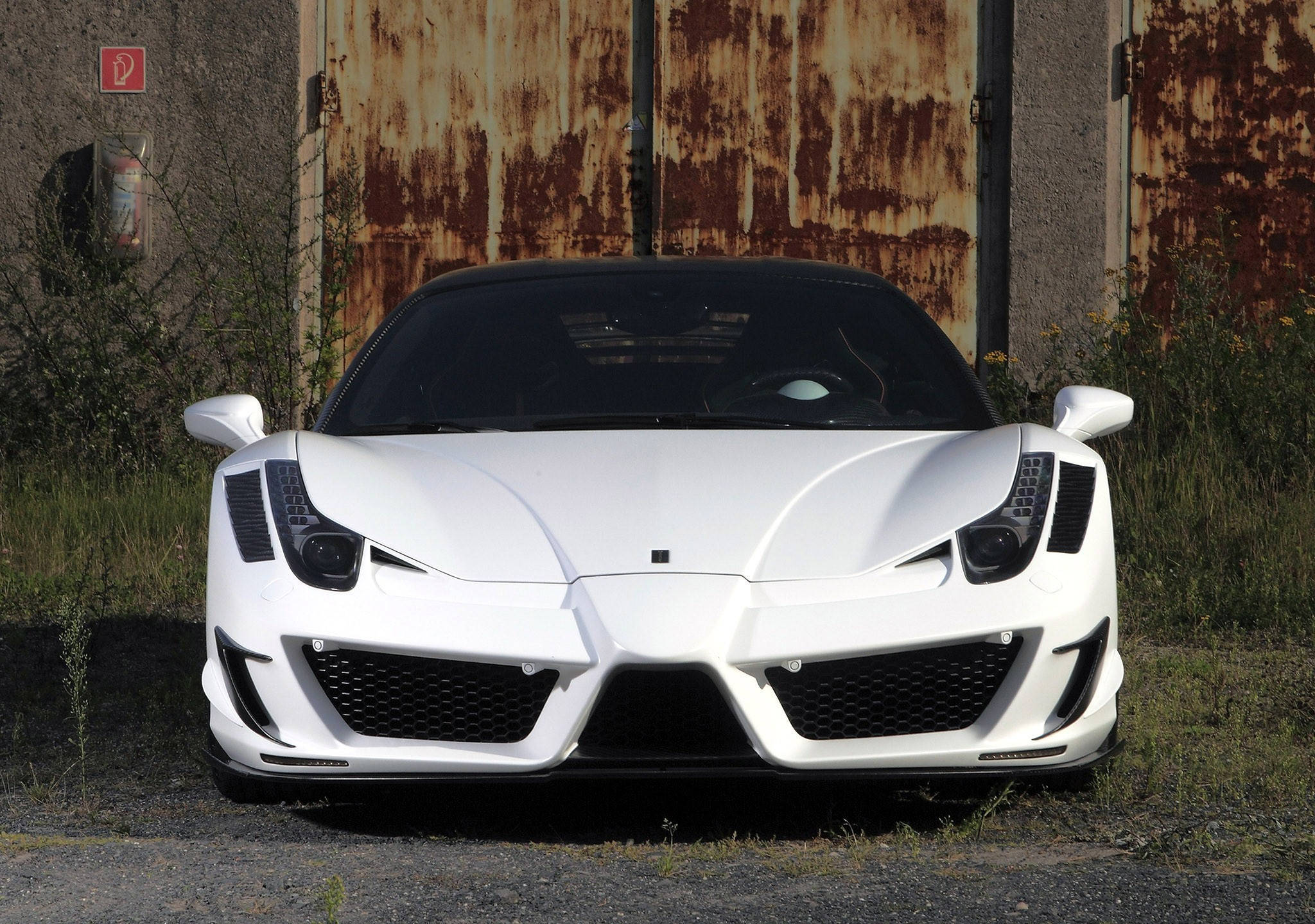 white ferrari 桌布 壁紙 — 一輛純白色的法拉利458 Italia正面朝前，停放在一扇風化生鏽的金屬門前，處於陽光普照的戶外環境中。