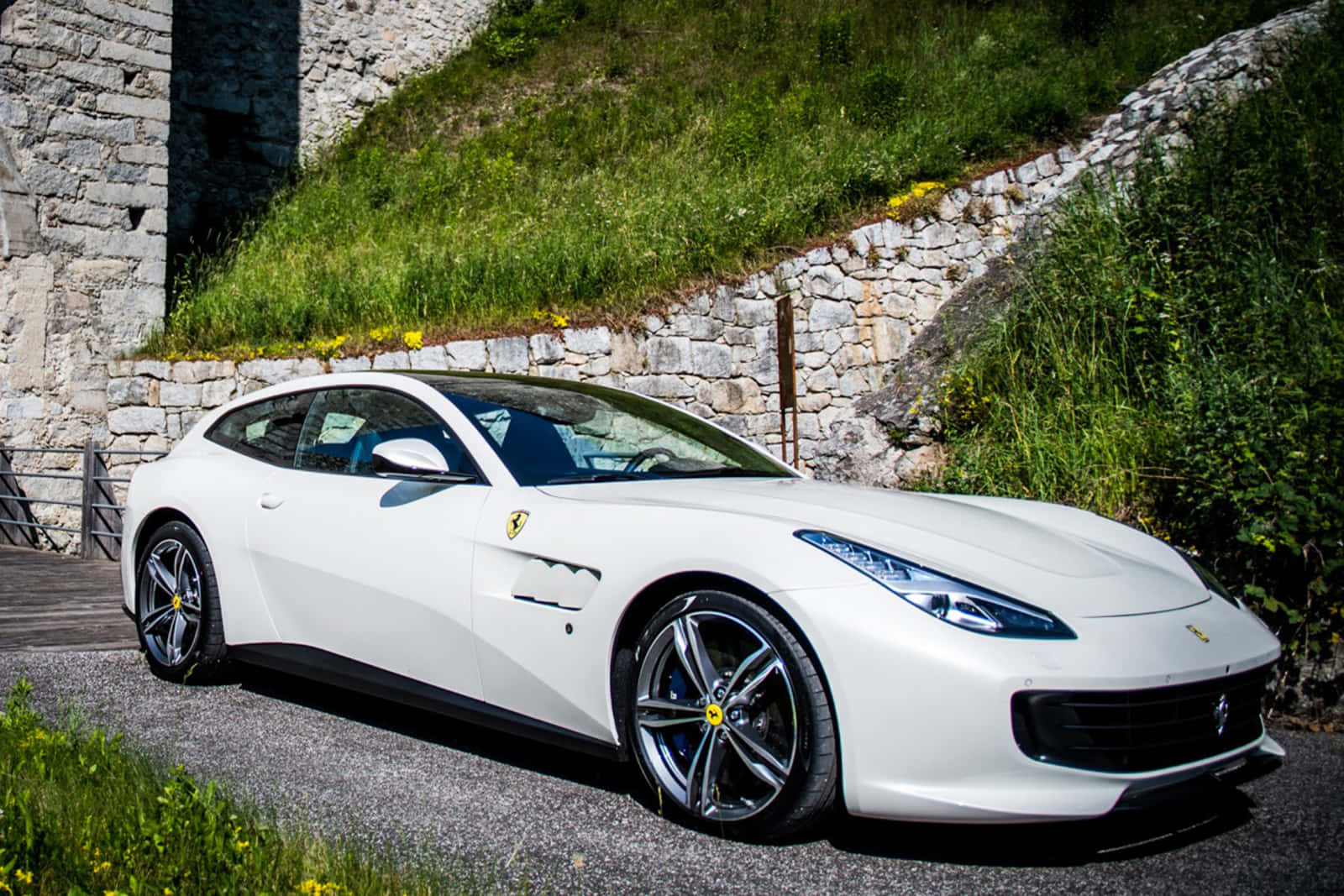 Ferrari Gtc4lusso 桌布 壁紙 — 一輛純白色的法拉利停泊在碎石路上，旁邊是石牆，有綠草和黃花。