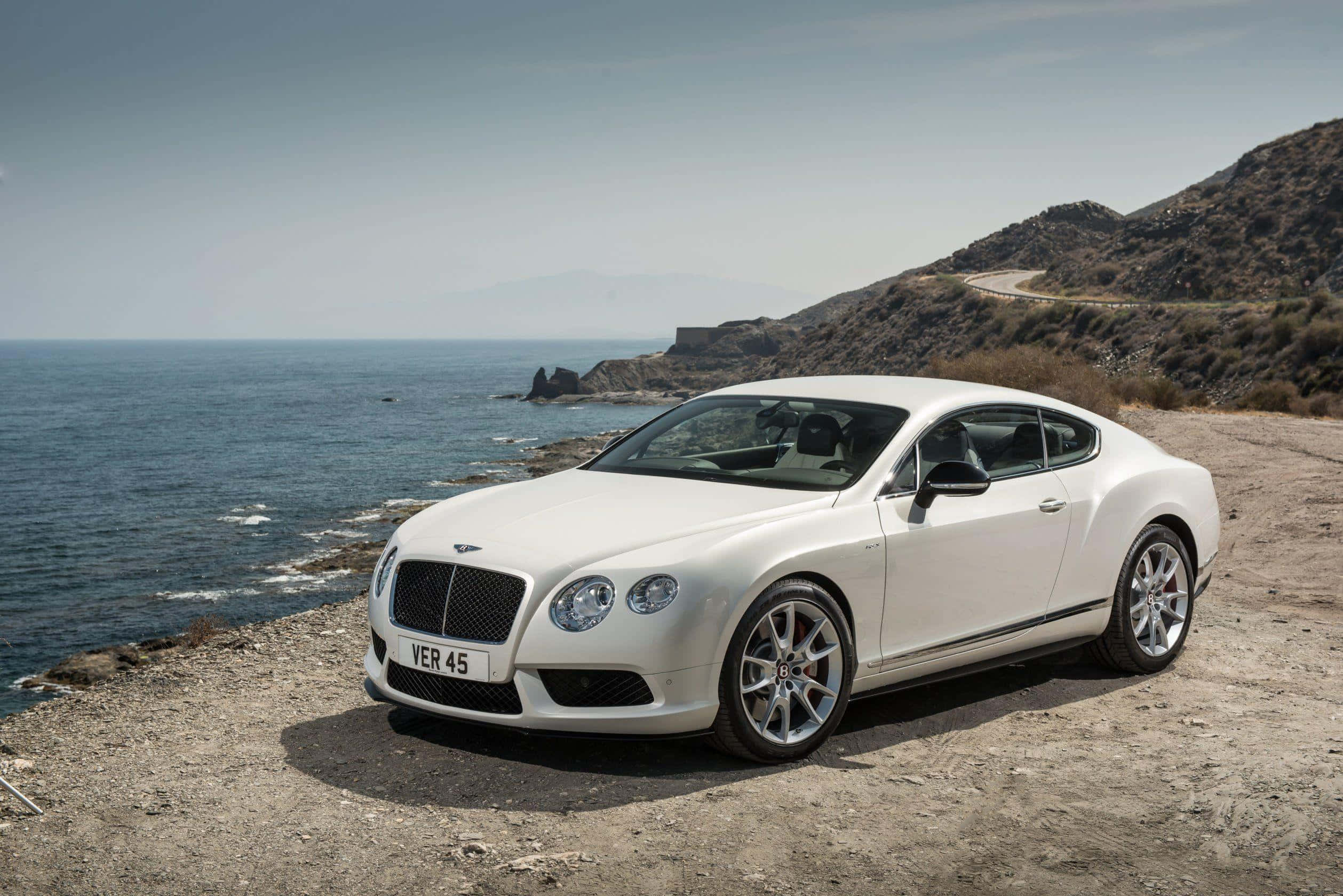 bentley HD 桌布 壁紙 — 一輛純白色的賓利 Continental GT 停泊在陽光明媚的懸崖上，俯瞰深藍色的海洋，背景是崎嶇的山丘。
