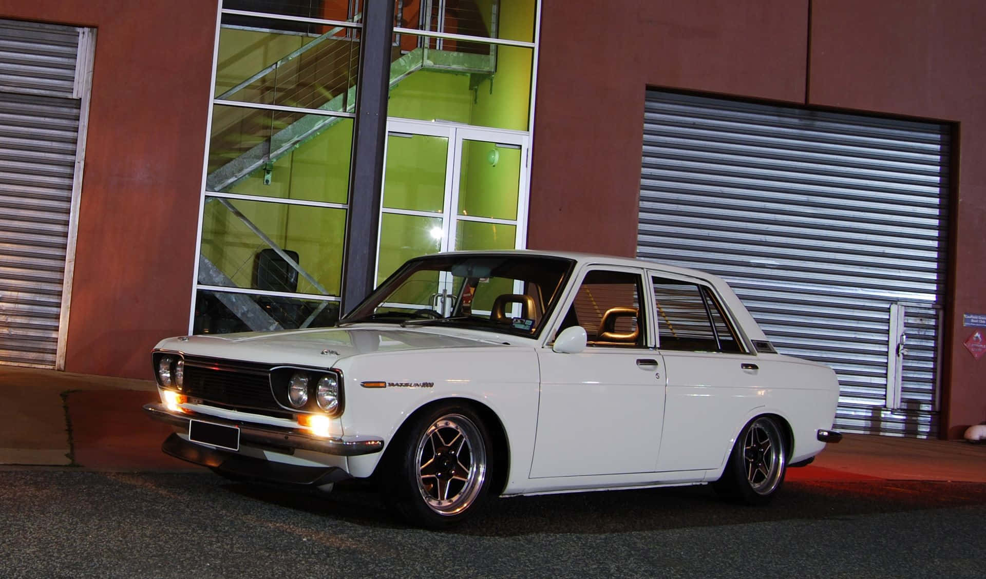 Datsun 桌布 壁紙 — 一輛純白色經典轎車配備訂製輪圈，在夜燈下閃閃發光，旁邊是一座帶有綠色發光窗戶的現代建築。