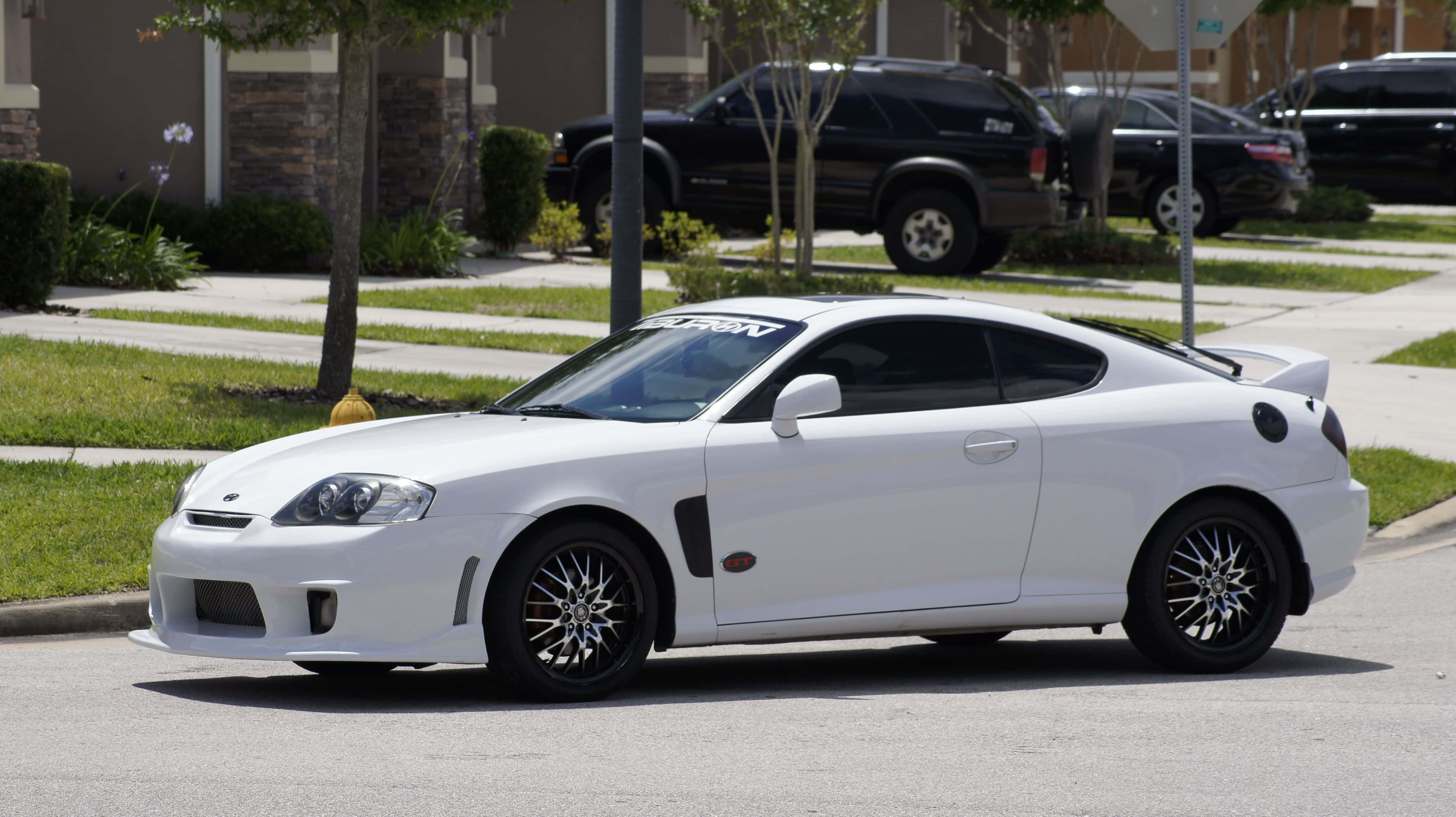 Hyundai Tiburon 桌布 壁紙 — 一輛純白色運動型轎跑車搭配黑色輪圈，停放在陽光明媚、綠草如茵、樹木成蔭的郊區街道上。