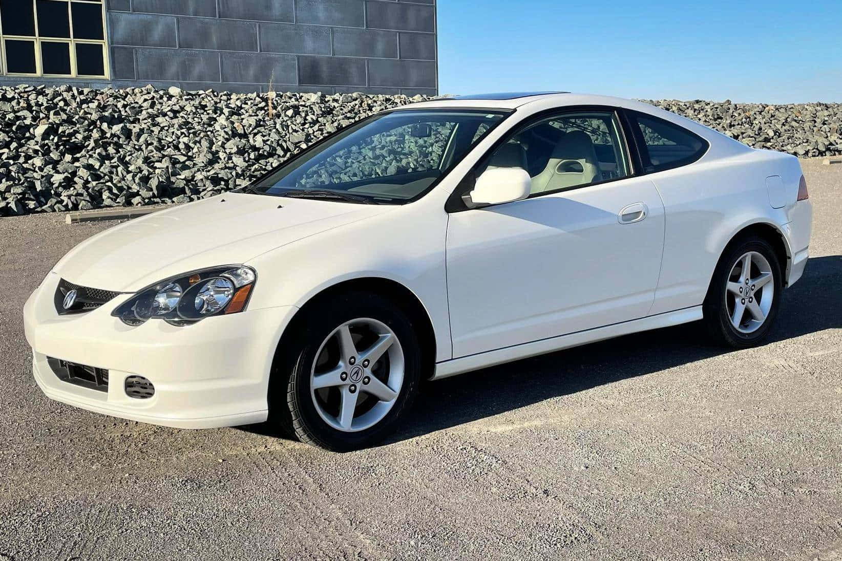 Acura Rsx 桌布 壁紙 — 一輛純白色雙門轎跑車停在碎石上，背景有石牆和灰色建築，天空湛藍清澈。