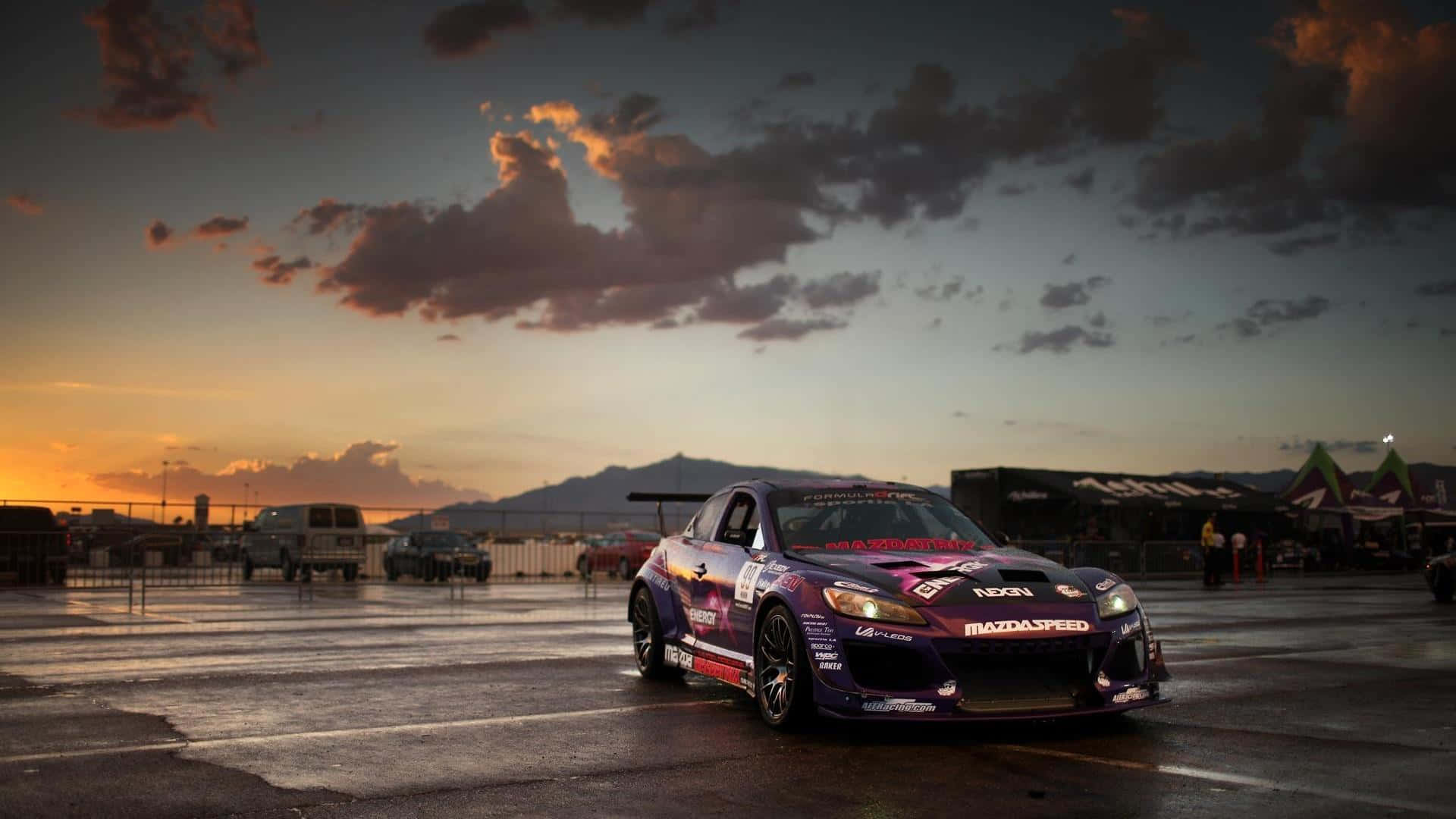 Mazda Rx-8 桌布 壁紙 — 一輛紫色Mazda飄移車在濕滑賽道上，於橙粉紅雲彩的戲劇性日落天空下閃耀。