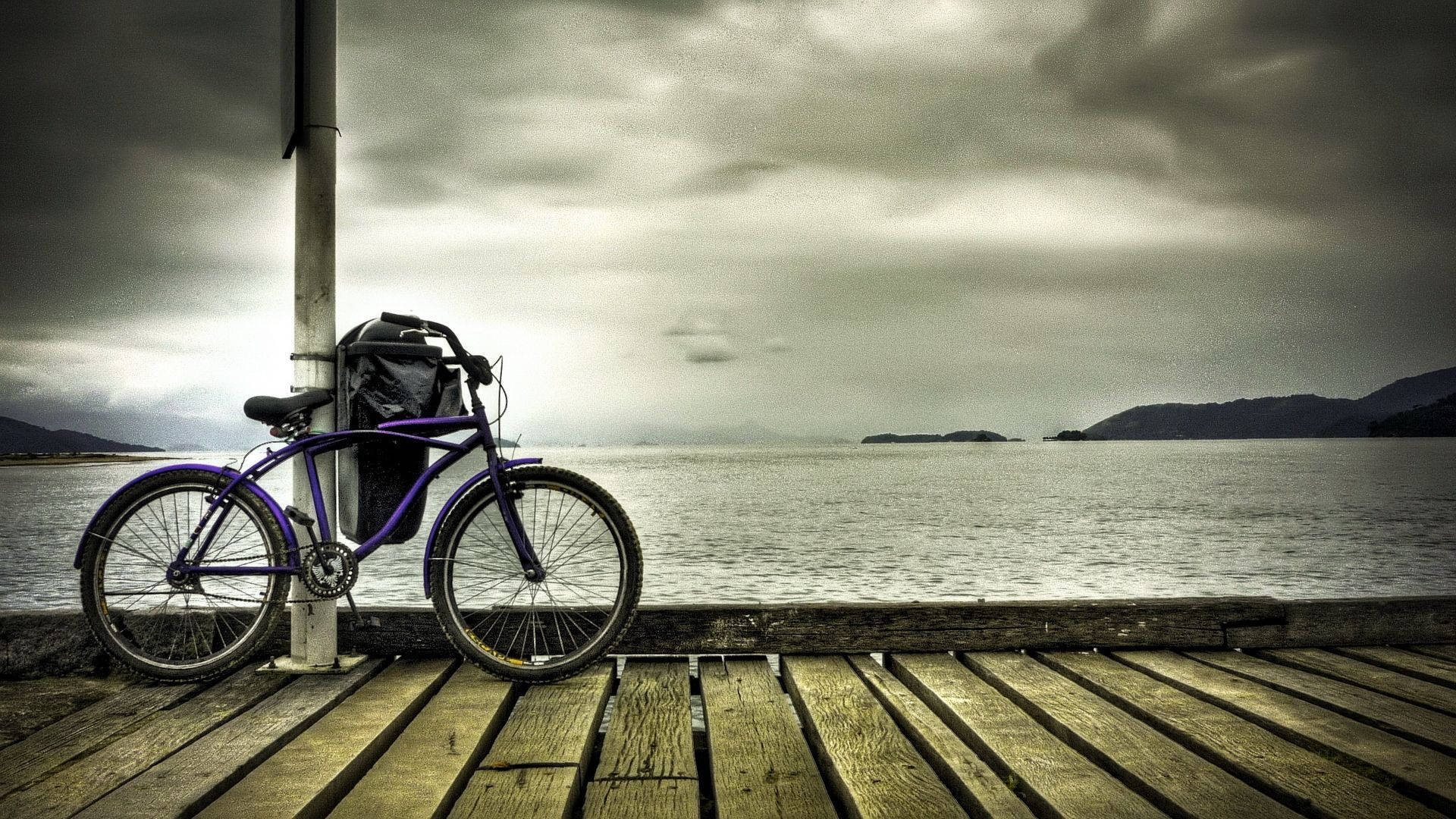 Bicycle 桌布 壁紙 — 一輛紫色腳踏車靠在一根柱子上，位於一個風化的木製碼頭，俯瞰著平靜的灰色海面，天空多雲。