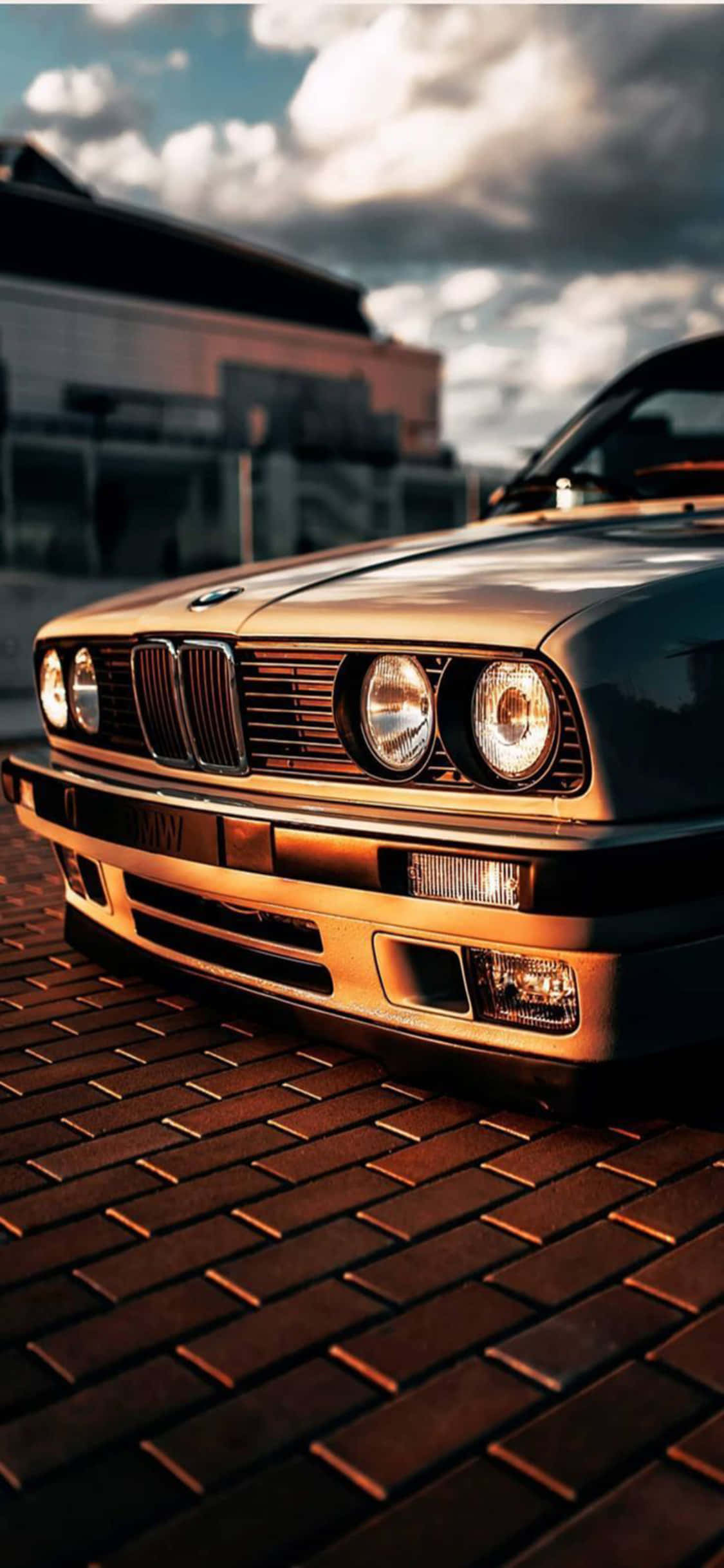 iPhone X Bmw 桌布 壁紙 — 一輛經典BMW E30跑車在金色夕陽下，於磚砌街道上閃耀，頭頂有戲劇性的雲層。