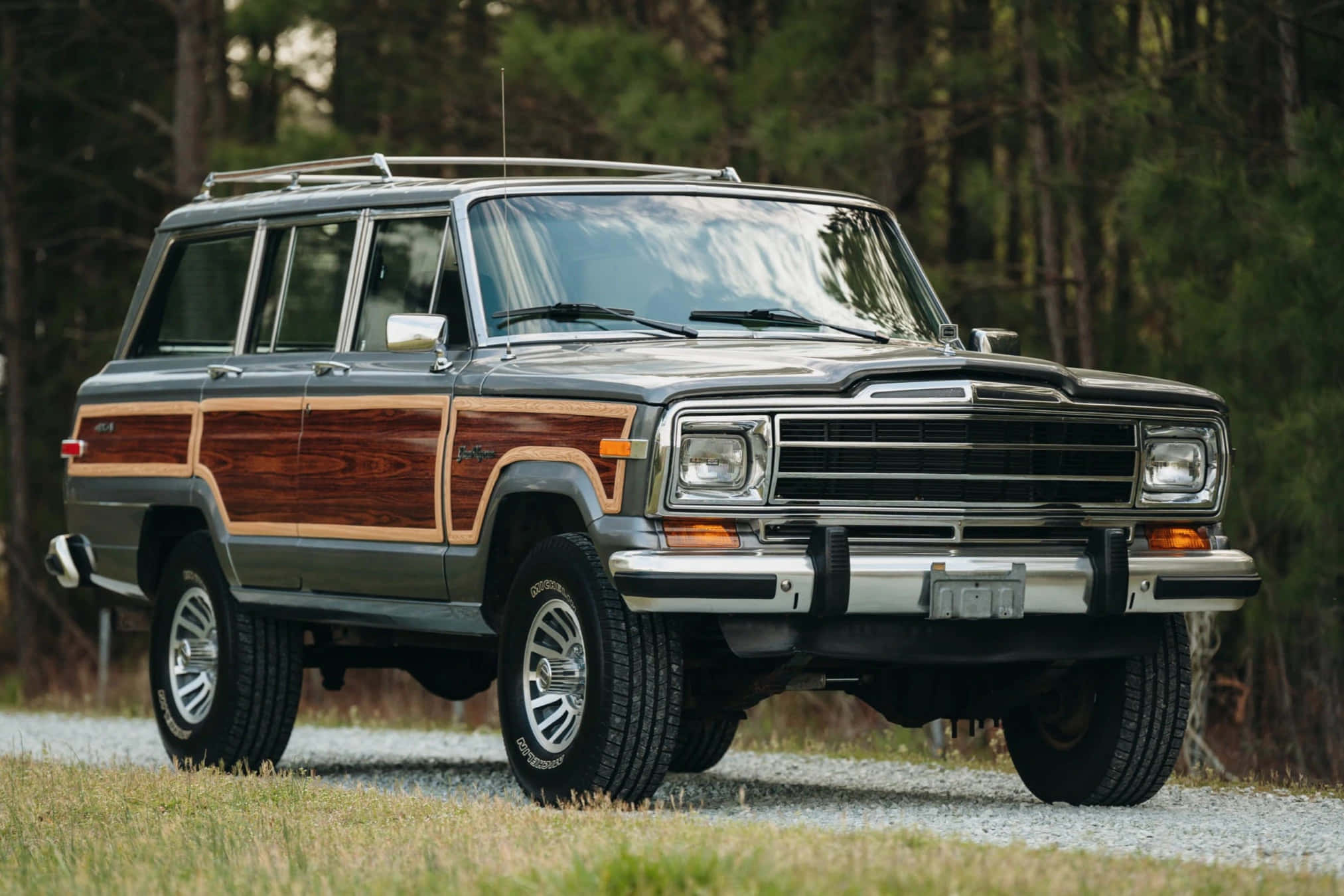 Jeep Wagoneer 桌布 壁紙 — 一輛經典木紋車身Jeep Cherokee旅行車停在碎石路上，周圍是高聳的松樹。