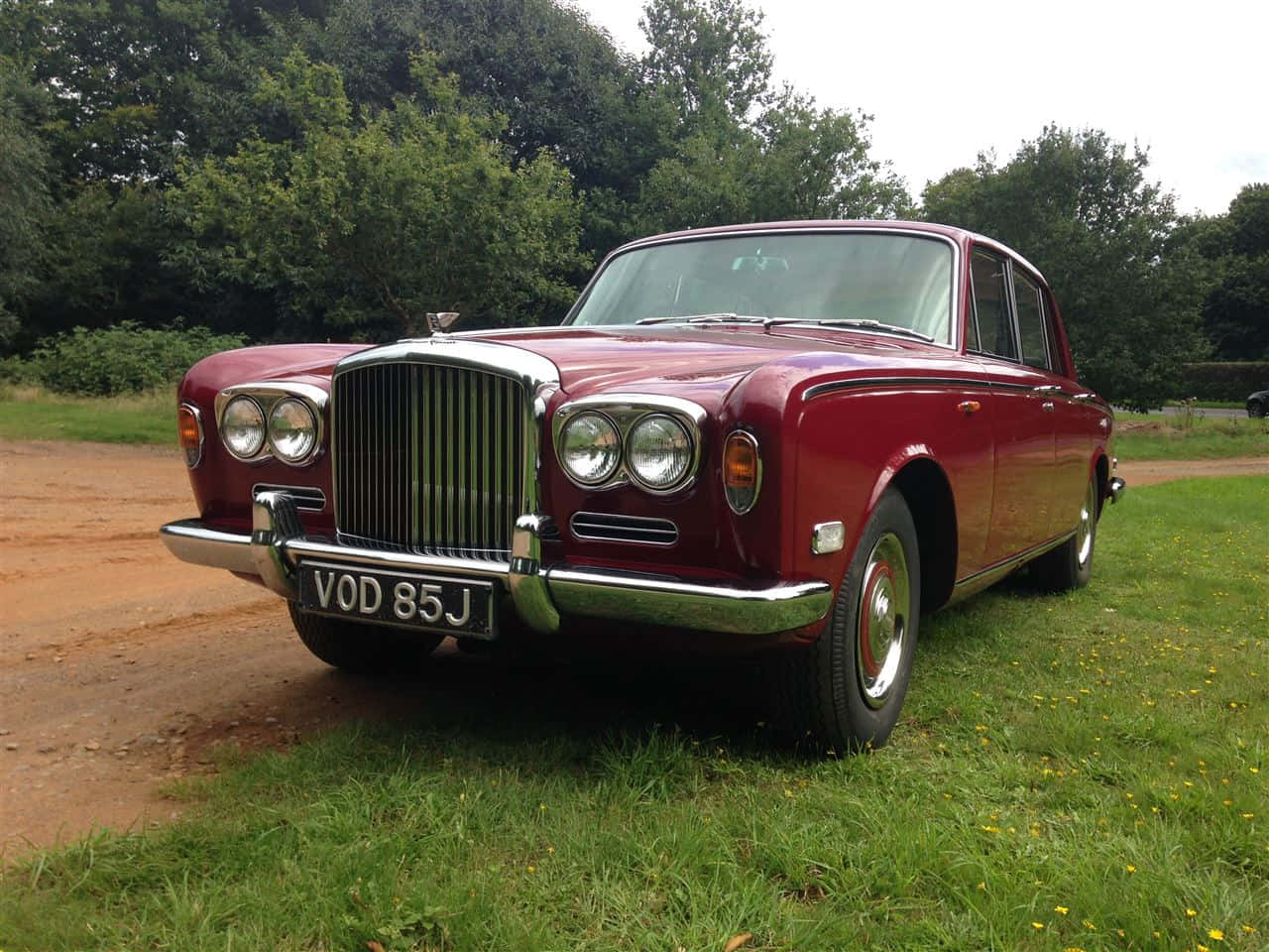 Bentley T1 桌布 壁紙 — 一輛經典栗色賓利轎車，帶有鍍鉻裝飾，停放在草地上，旁邊是一條泥濘小徑，天空多雲。