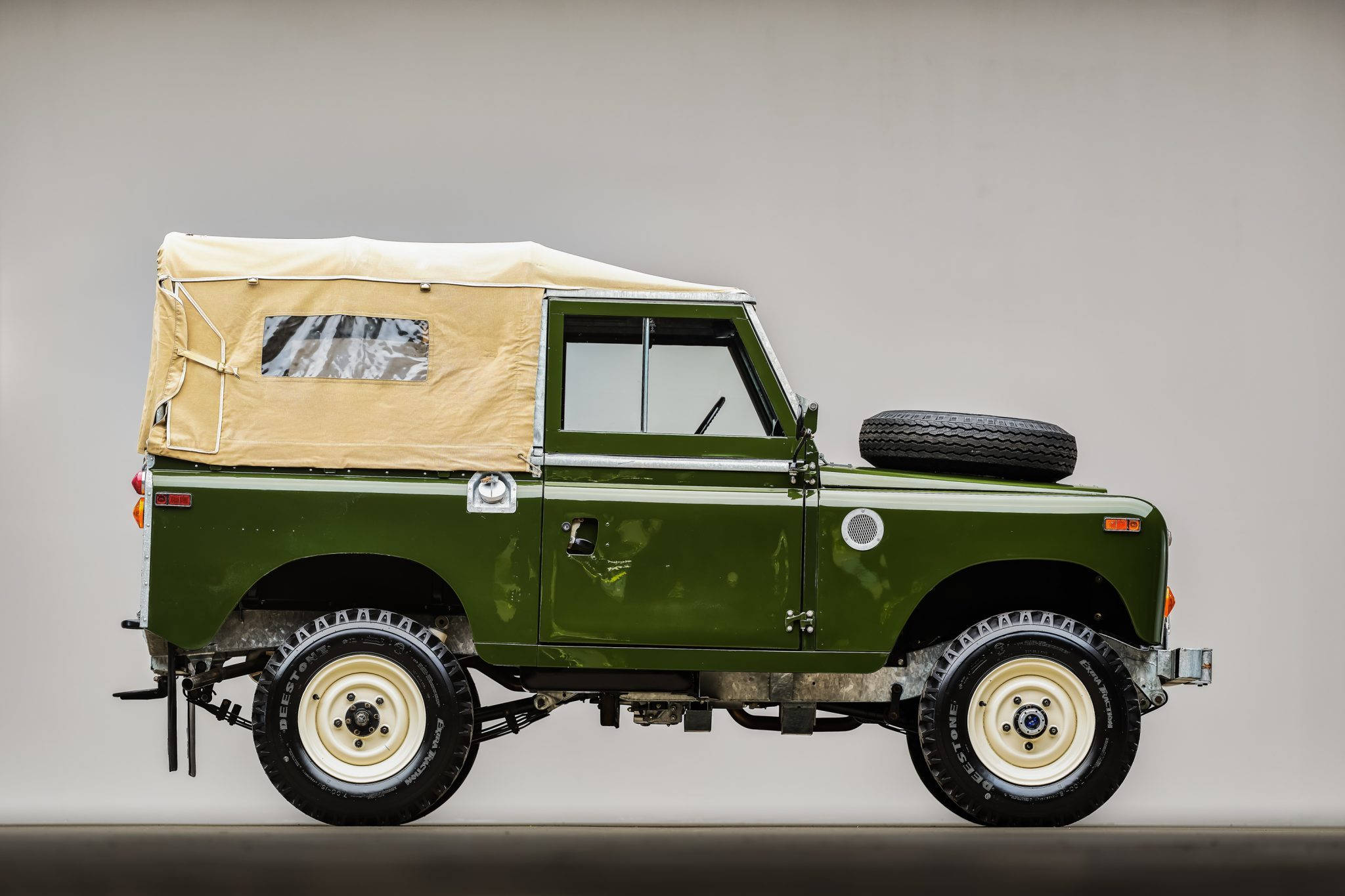 land rover iPhone 桌布 壁紙 — 一輛經典橄欖綠色Land Rover，配有米色帆布車頂和安裝在引擎蓋上的備用輪胎，矗立在攝影棚環境中。