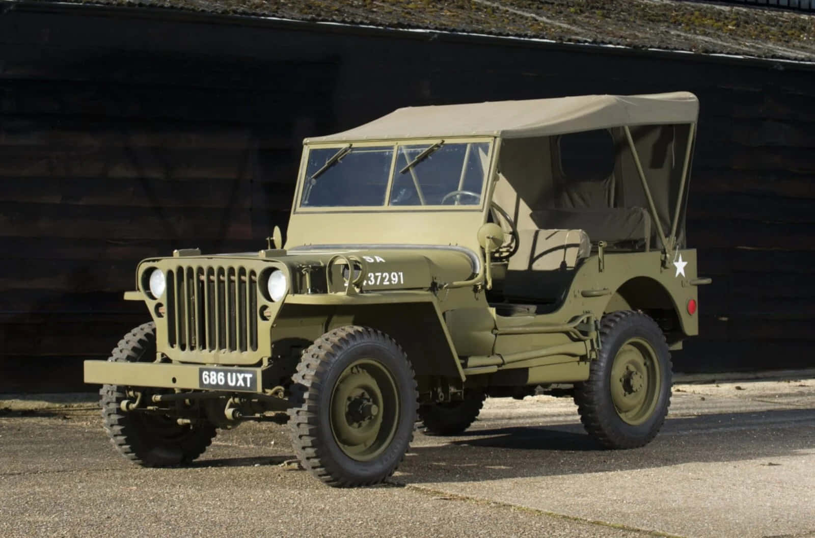 Jeep Willys 桌布 壁紙 — 一輛經典橄欖綠軍用吉普車，帶帆布頂篷，停在柏油路面上，背景是深色木牆。