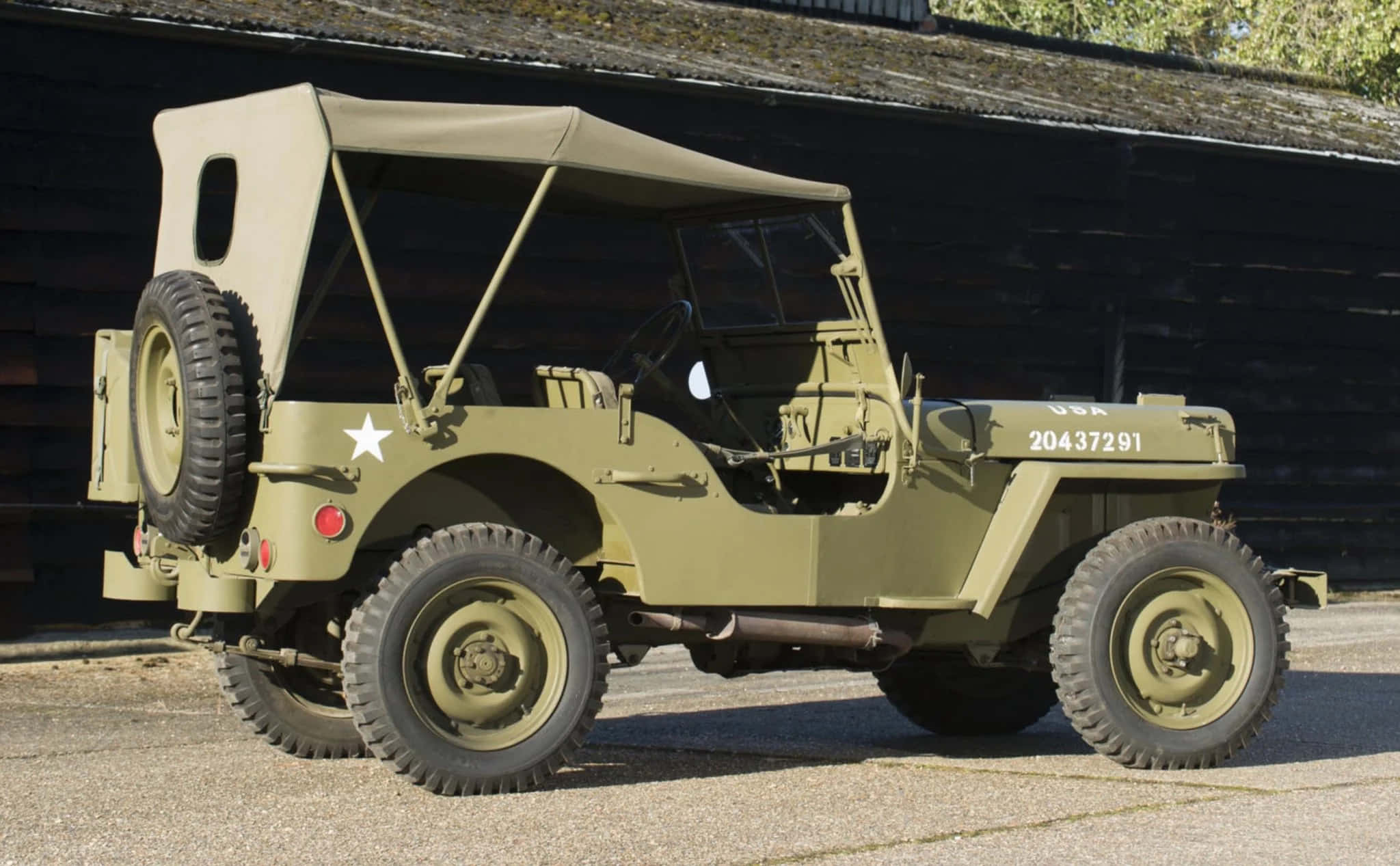 Jeep Willys 桌布 壁紙 — 一輛經典橄欖綠軍用吉普車，帶有白色星星標誌，停在木棚旁邊的礫石上，光線明亮。