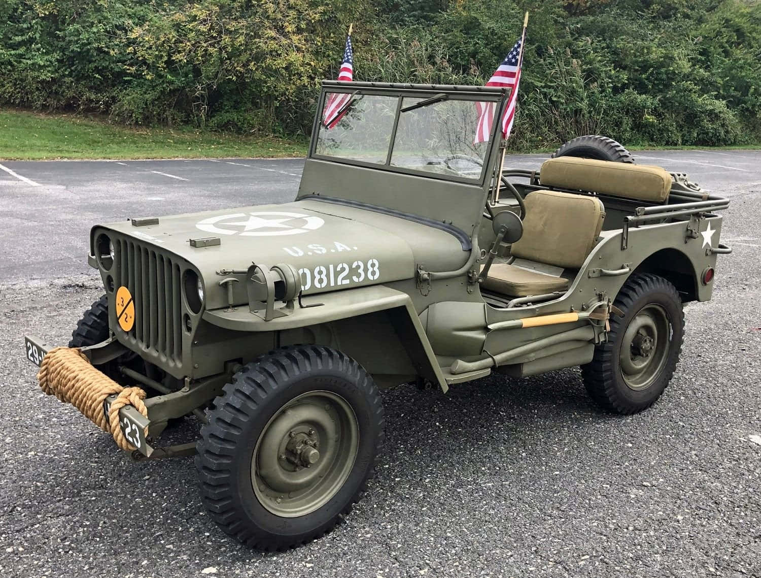 Jeep Willys 桌布 壁紙 — 一輛經典橄欖綠軍用吉普車，掛有美國國旗，停泊在碎石地上，四周綠葉環繞。