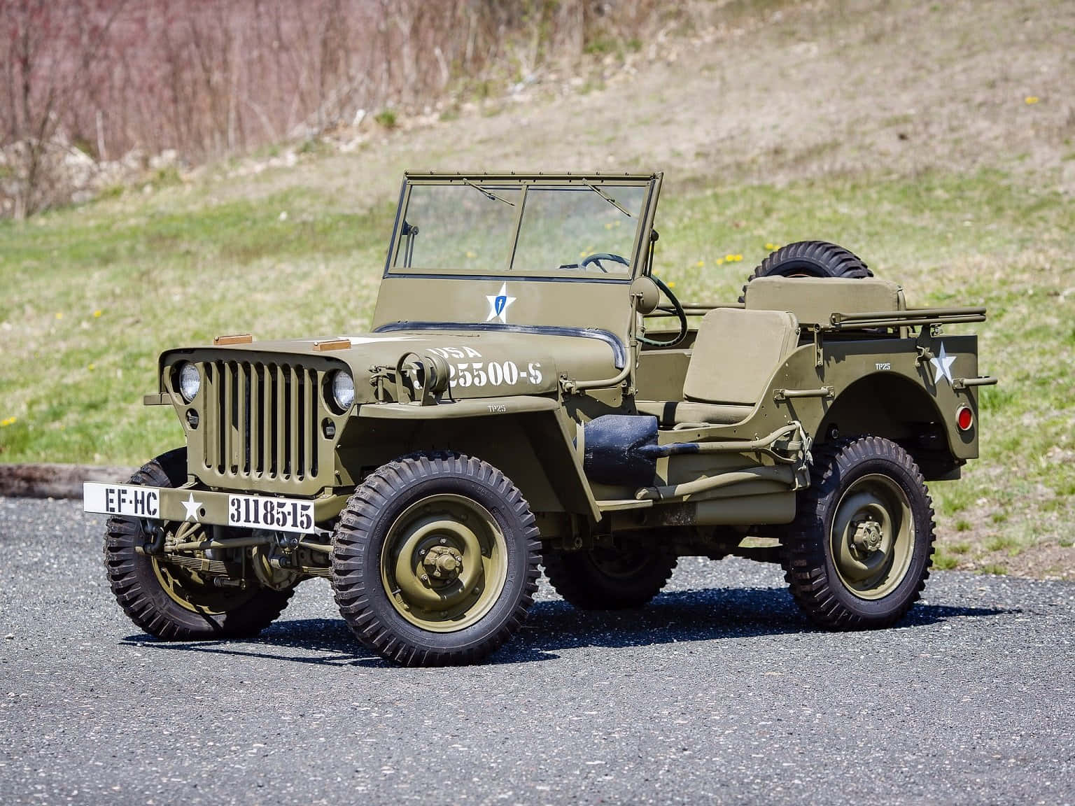 Jeep Willys 桌布 壁紙 — 一輛經典橄欖綠軍用吉普車帶有星形徽章，在明亮的日光下停泊在碎石路上，旁邊是草地地形。