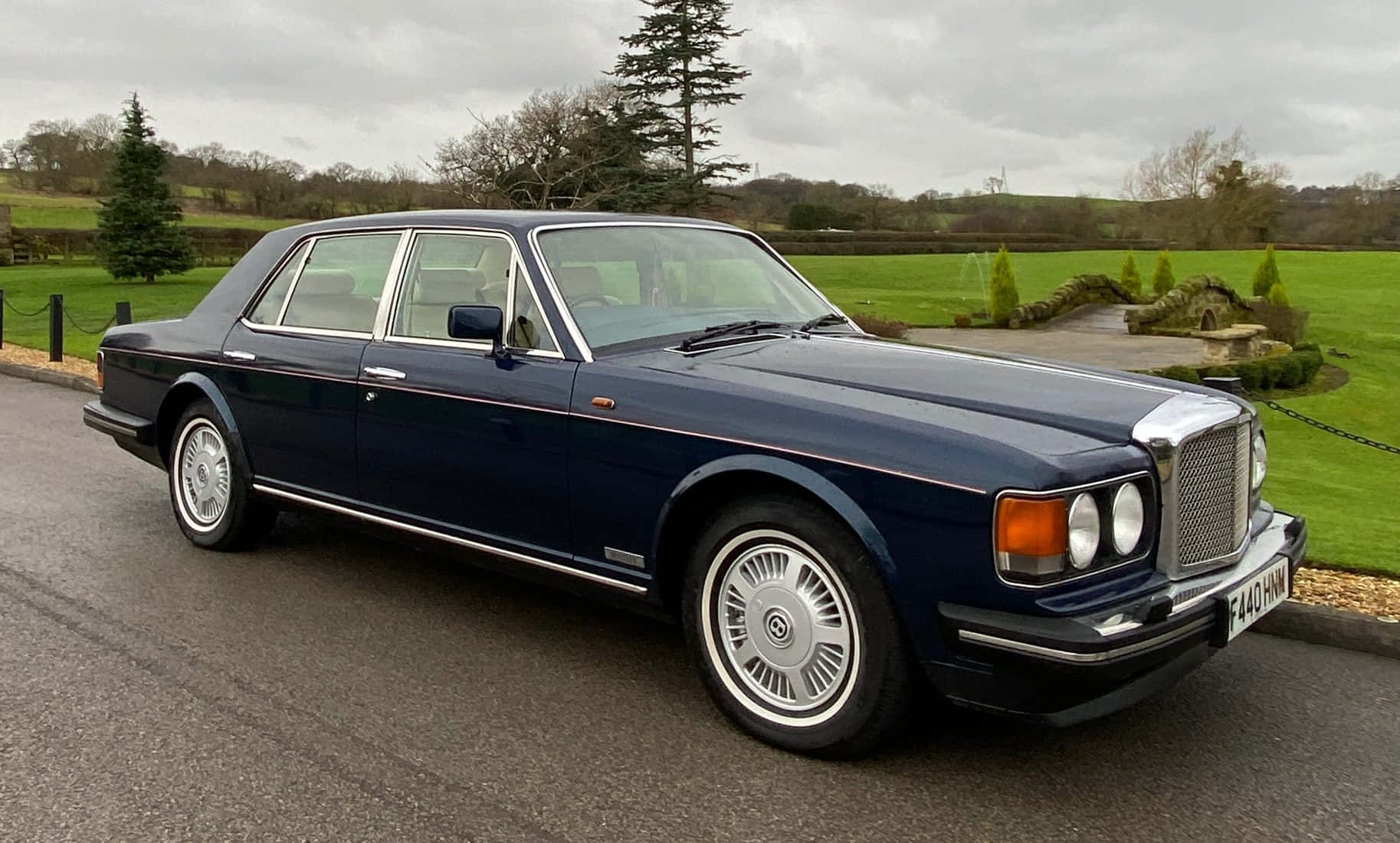 Bentley Eight 桌布 壁紙 — 一輛經典海軍藍色賓利轎車在多雲的天空下閃閃發光，停在鋪砌的車道上，旁邊是修剪整齊的綠色草坪和石頭花園特色。