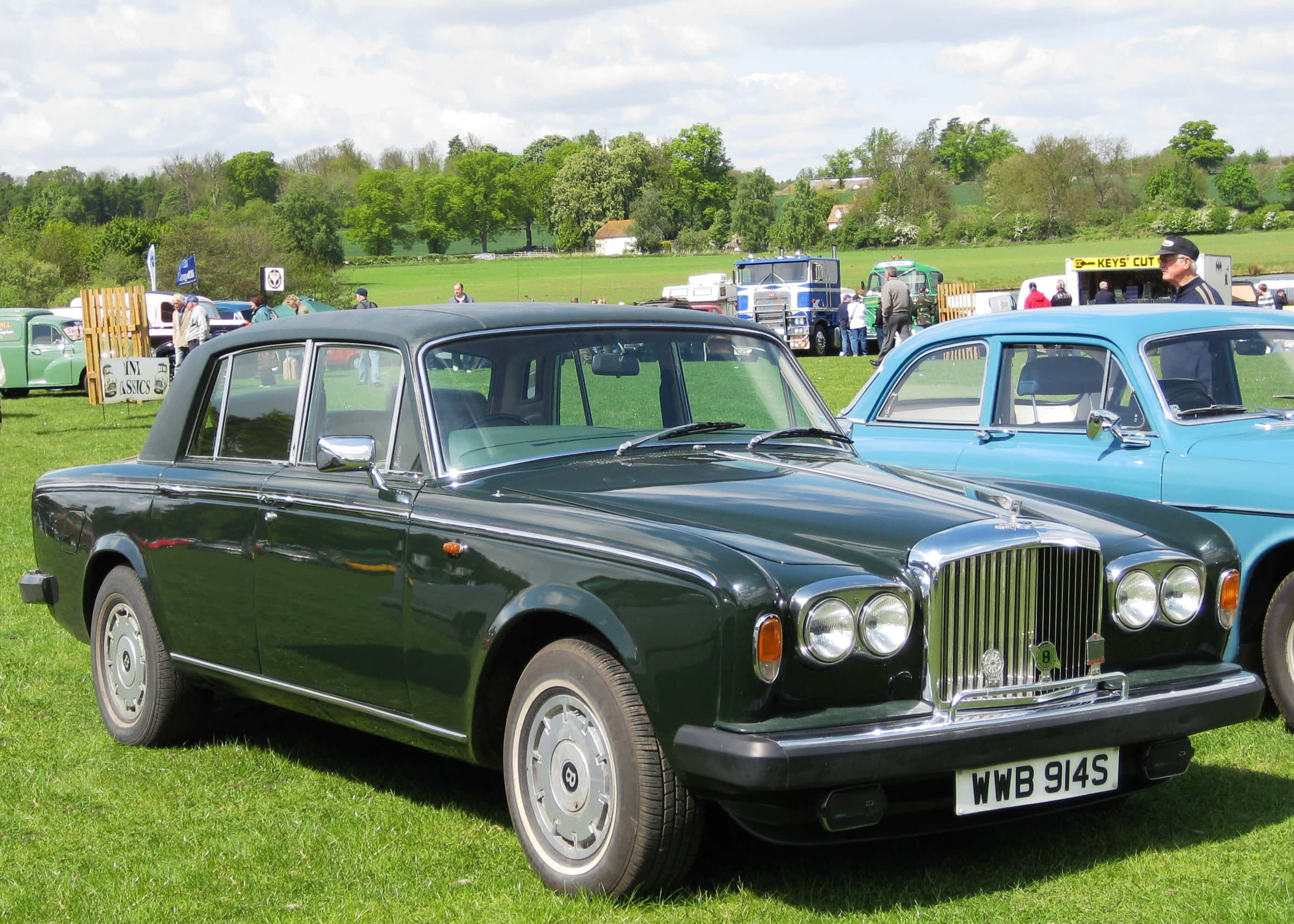 Bentley T2 桌布 壁紙 — 一輛經典深綠色賓利轎車在明媚陽光下，於經典車展的草地上閃閃發光，旁邊有一輛藍色汽車。