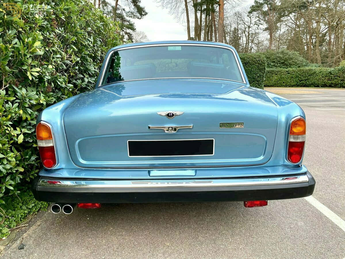 Bentley T2 桌布 壁紙 — 一輛經典淺藍色 Bentley T2 停泊在柏油路上，旁邊有綠色籬笆，天空多雲。