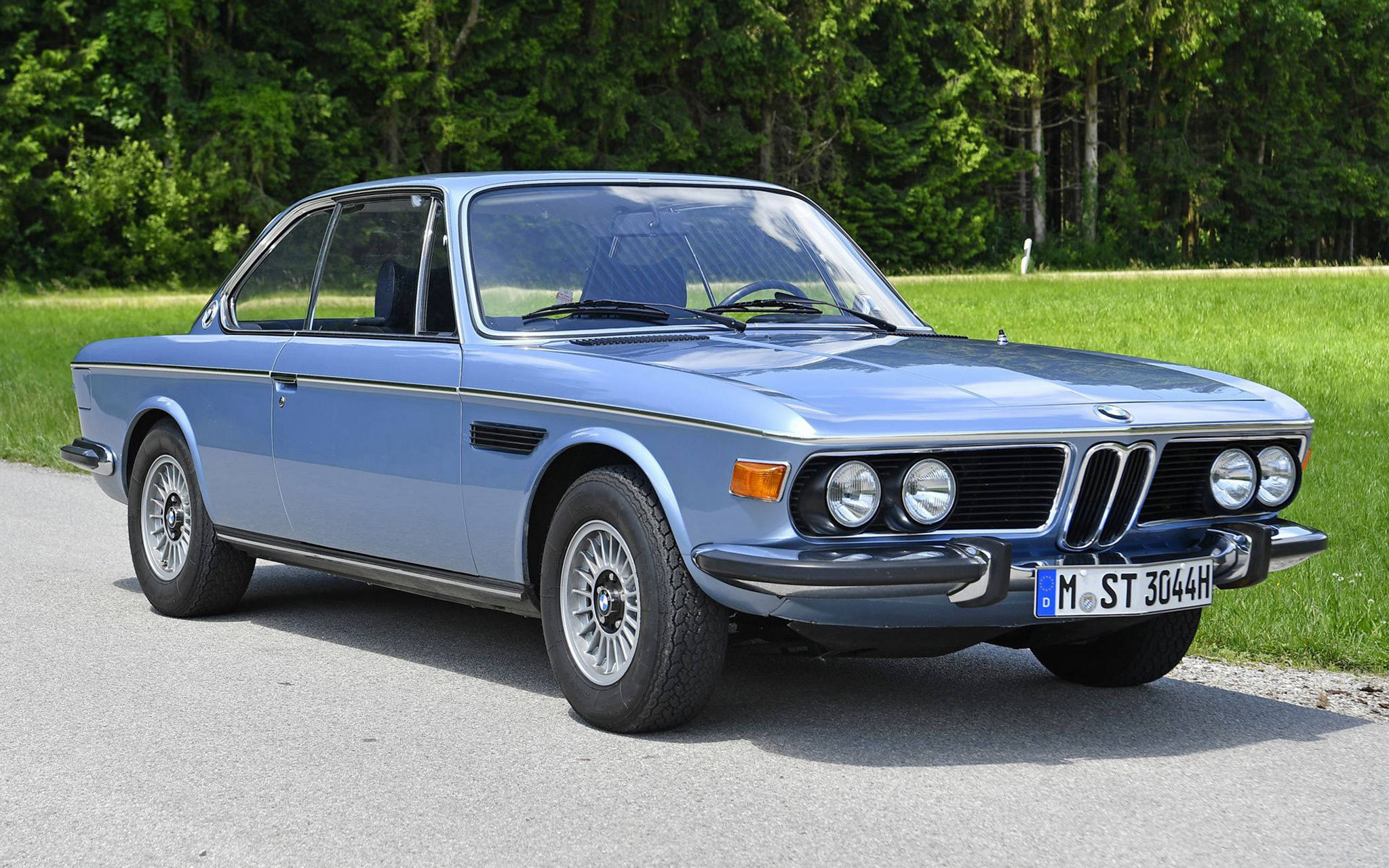classic bmw 桌布 壁紙 — 一輛經典淺藍色BMW 3.0 CSL雙門跑車停泊在鋪砌道路上，旁邊是茂密的綠色田野和濃密的森林。