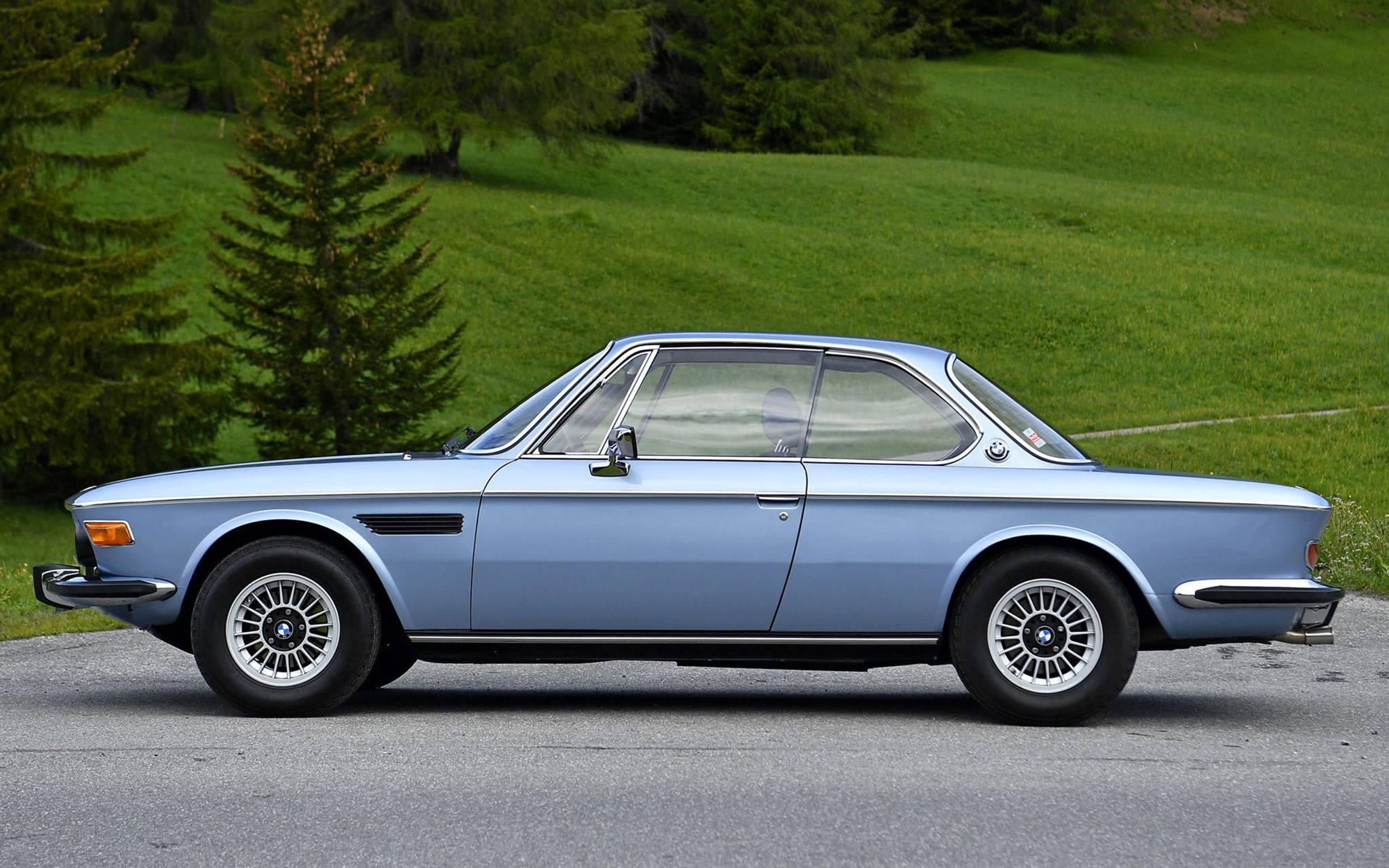 classic bmw 桌布 壁紙 — 一輛經典淺藍色BMW雙門跑車停放在鋪砌道路上，背景是茂密的綠色山丘和常綠樹木。