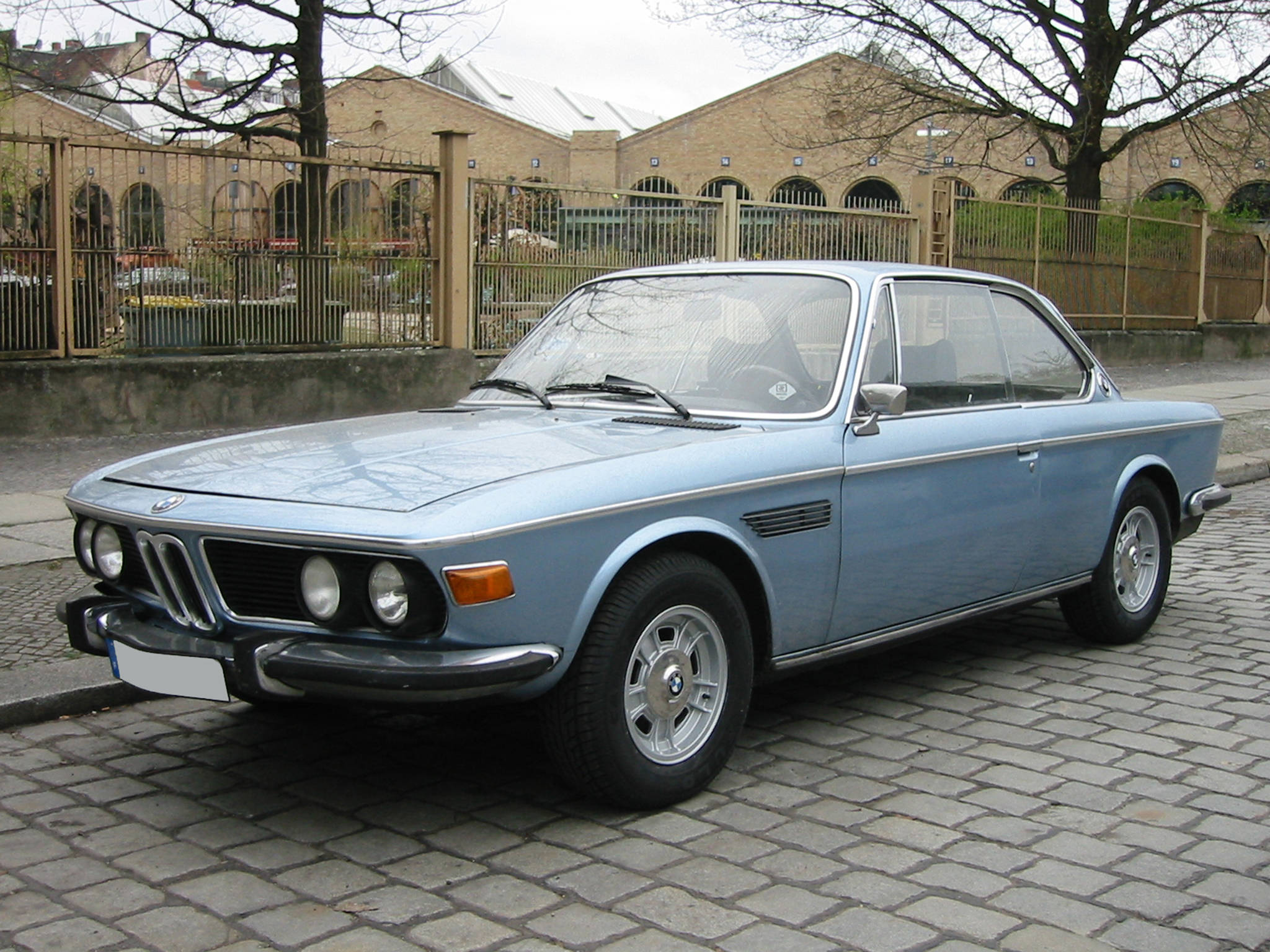 classic bmw 桌布 壁紙 — 一輛經典淺藍色寶馬轎跑車停泊在鵝卵石街道上，背景有光禿的樹木和磚砌建築。
