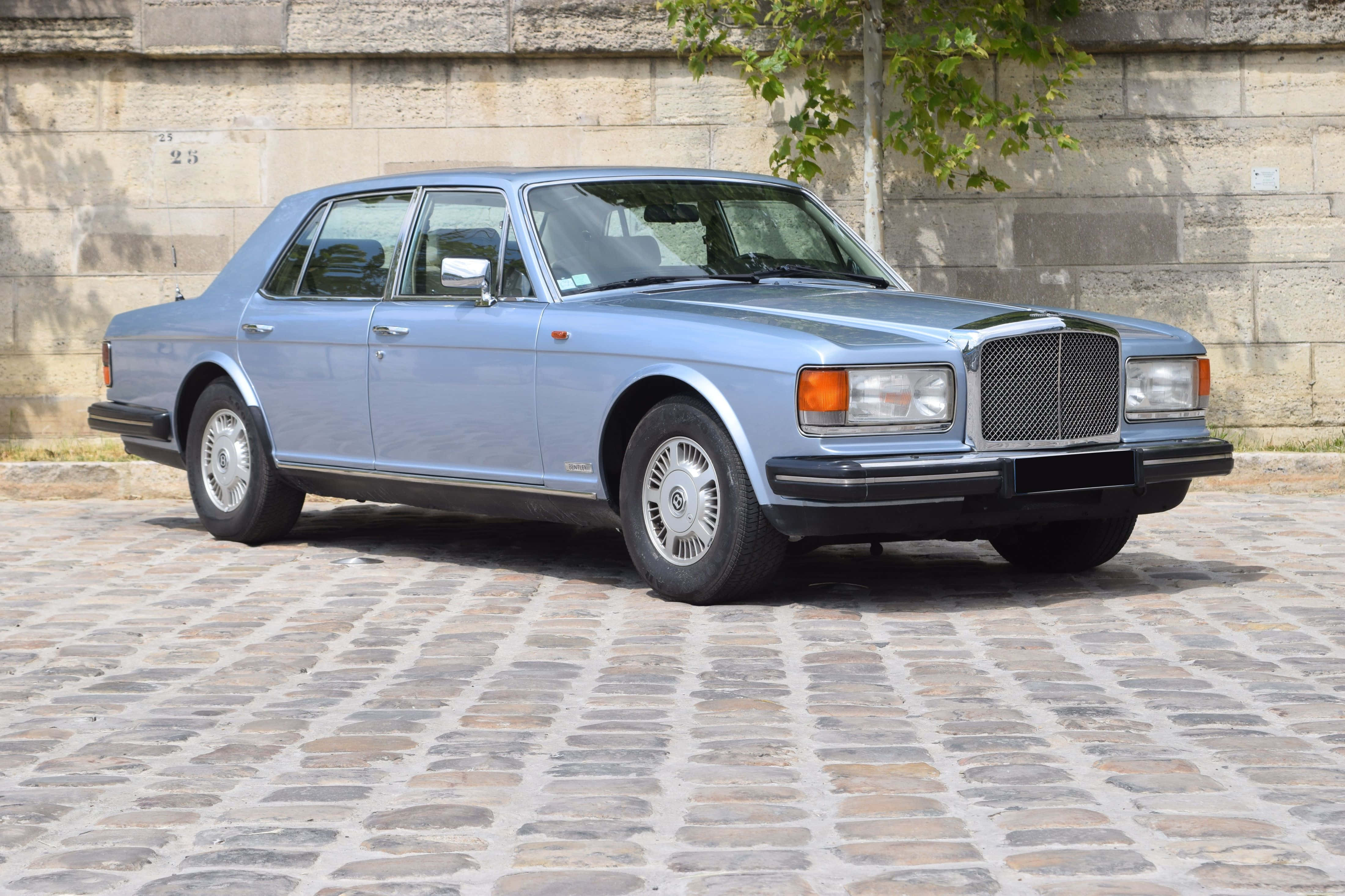 Bentley Eight 桌布 壁紙 — 一輛經典淺藍色賓利房車停泊在陽光普照的鵝卵石街道上，旁邊是帶有綠色植物的石牆。