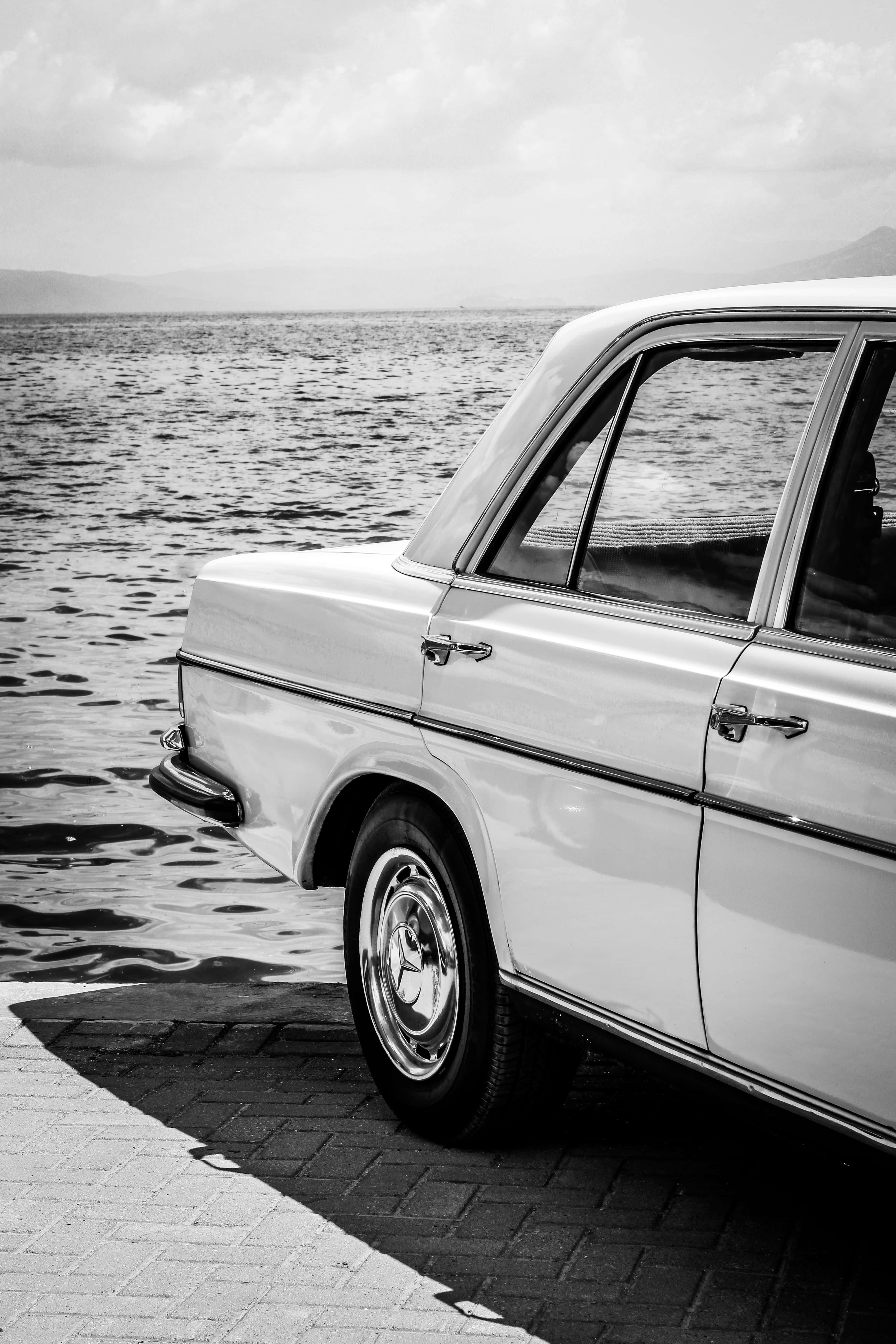 Classic Mercedes 桌布 壁紙 — 一輛經典白色轎車停泊在磚砌碼頭上，海浪拍打輪胎，天空多雲。